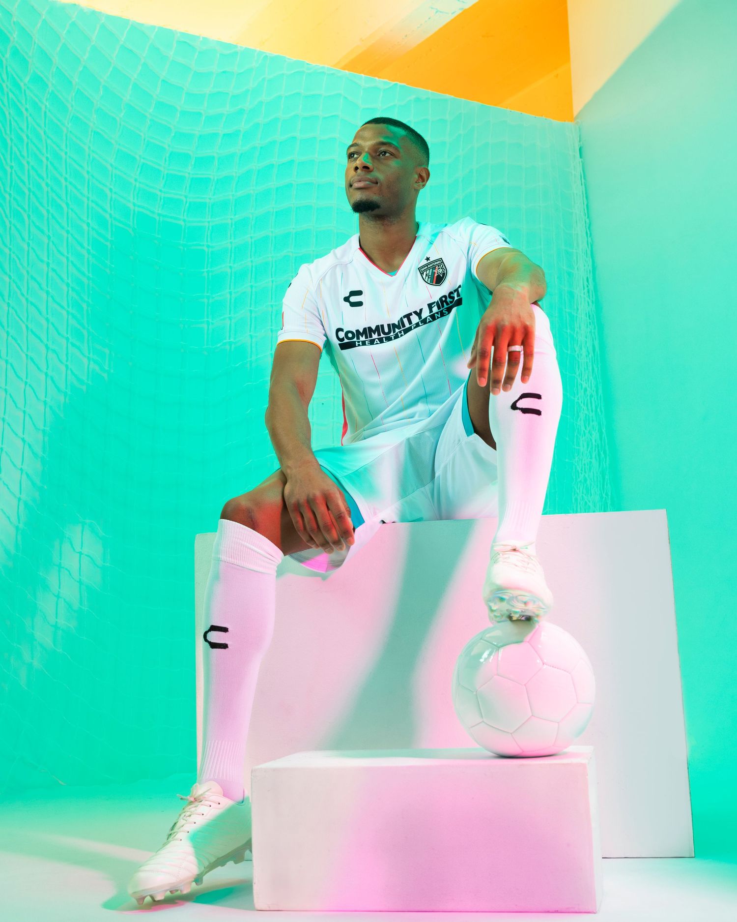 Soccer player in white uniform sits on geometric blocks against turquoise and pink background.
