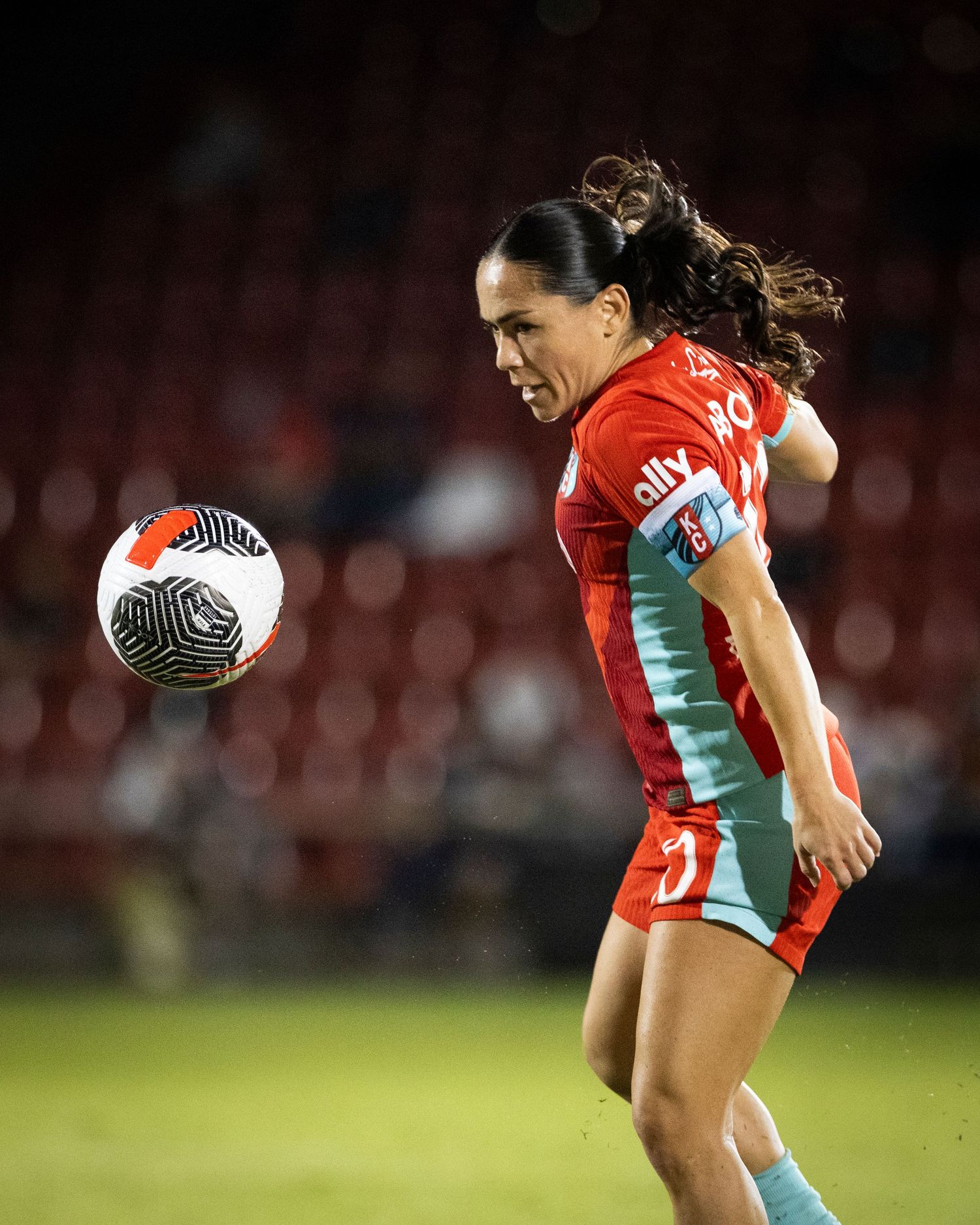 Soccer player controls ball during night game wearing red and turquoise uniform.