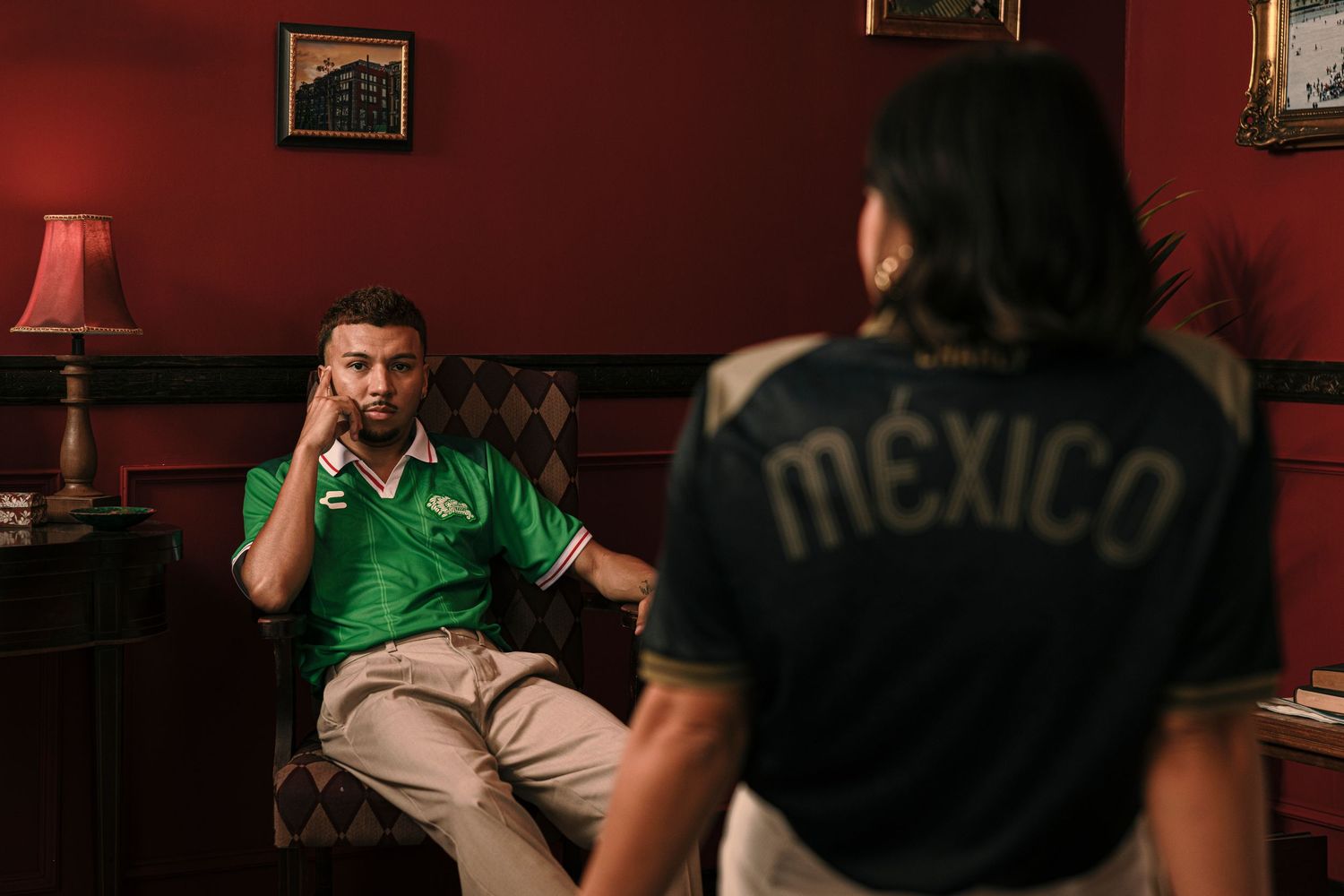 Two men in a red room, one wearing a green polo and one in a Mexico soccer jersey, having a conversation.