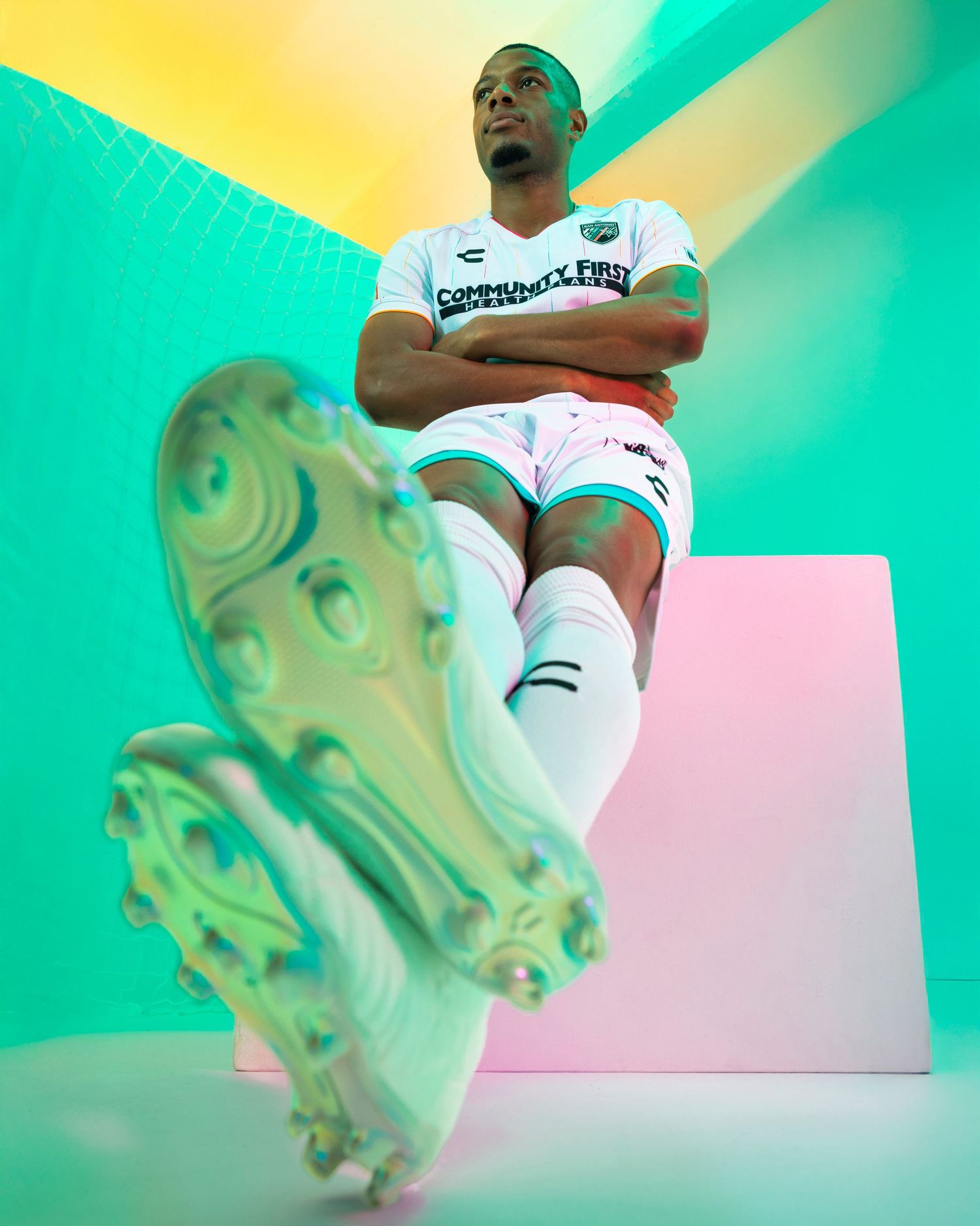 Soccer player in white uniform shows cleats while posing against vibrant geometric background.