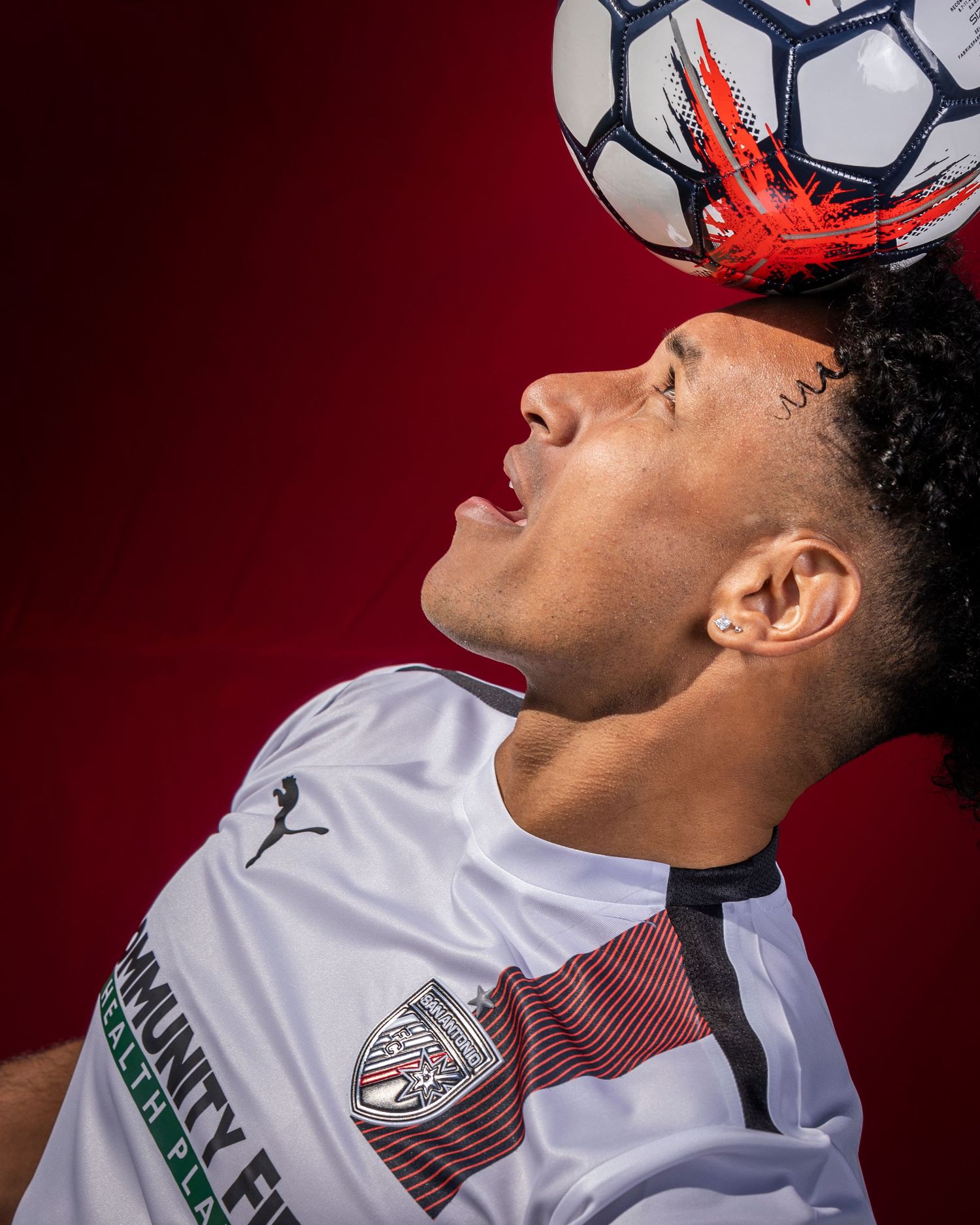 Close-up of player balancing soccer ball on head against red backdrop wearing white uniform.