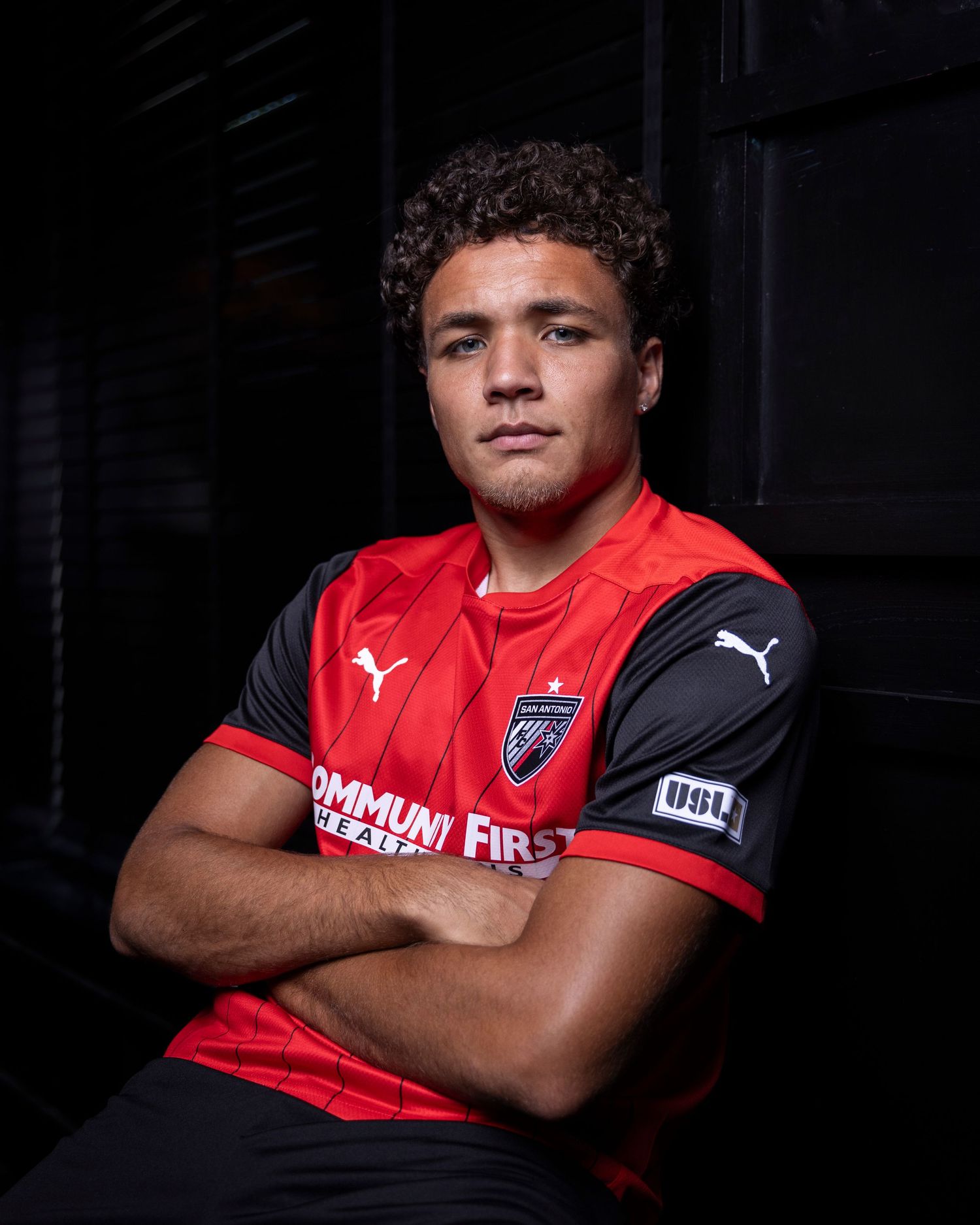 Soccer athlete in red and black Puma jersey poses with arms crossed in dramatic lighting.