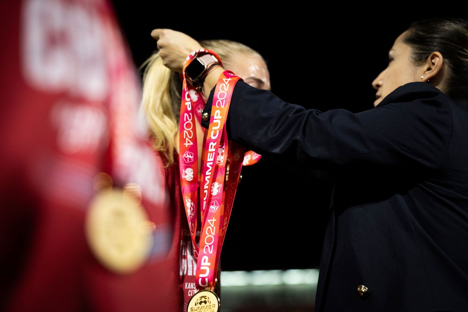 Hands present medals during awards ceremony with red ribbon against dark background.