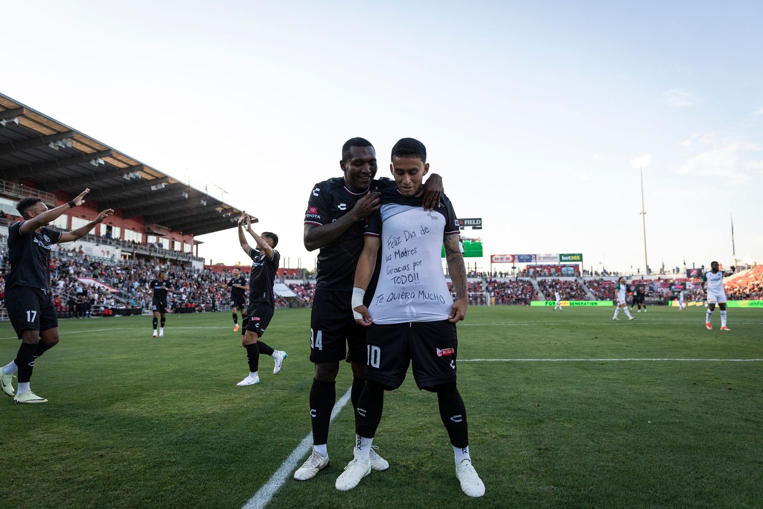 Athletes stand on a soccer field during a match with stadium seating visible in background.