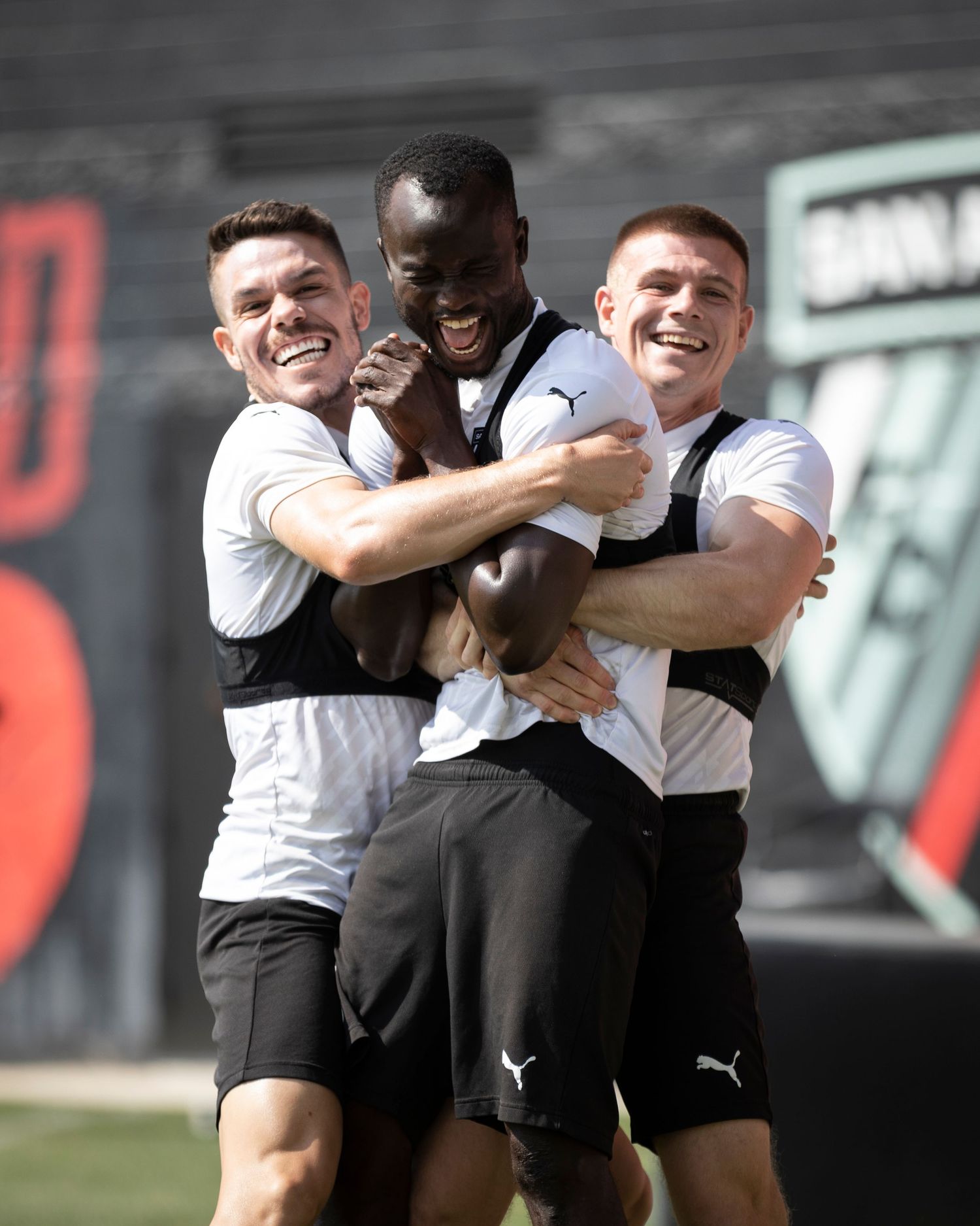 Three teammates share joyful embrace during training session in black and white uniforms.