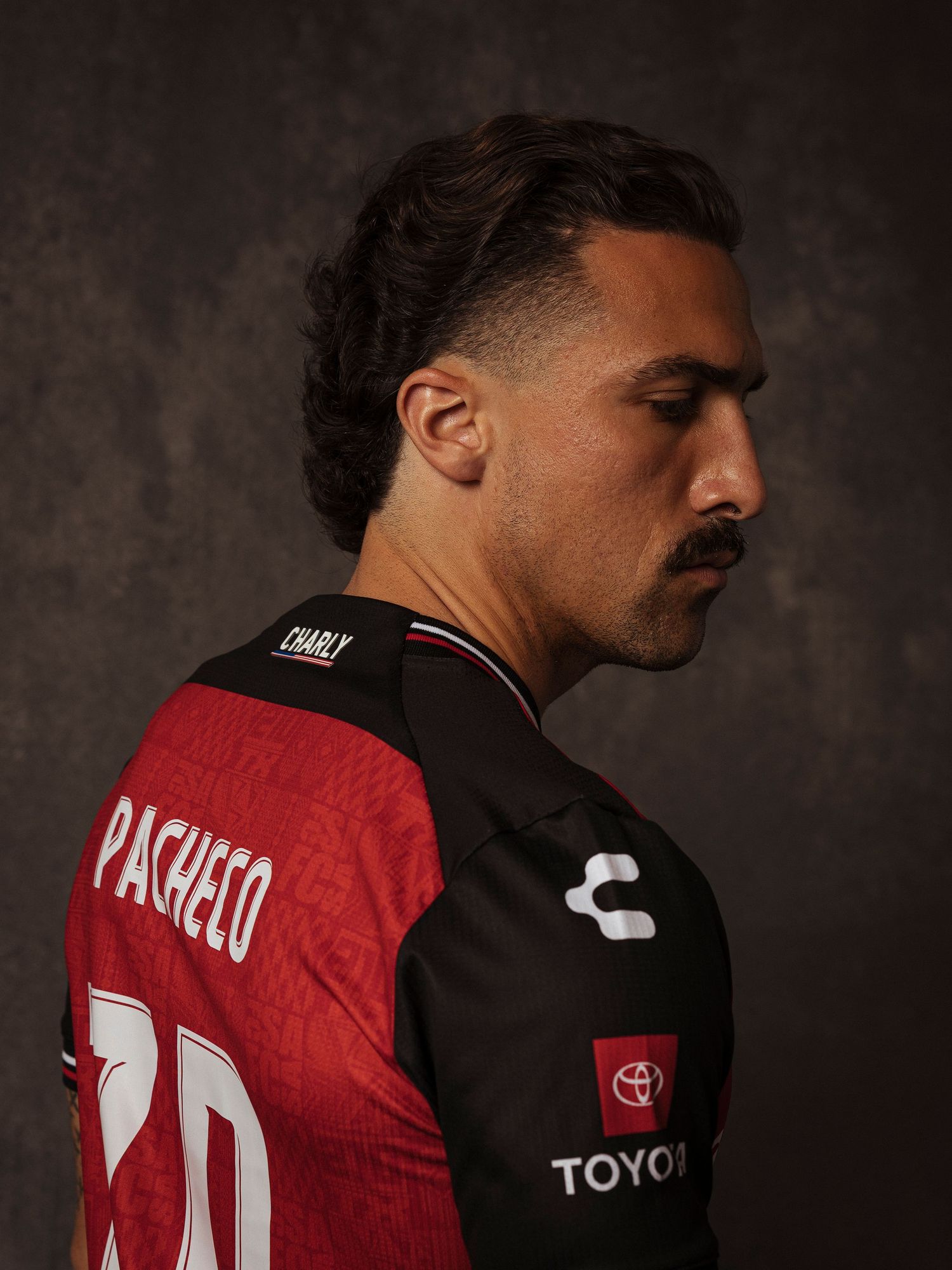Soccer player wearing a Charly brand jersey with PACHECO name and Toyota sponsor, posing in a dark studio setting.