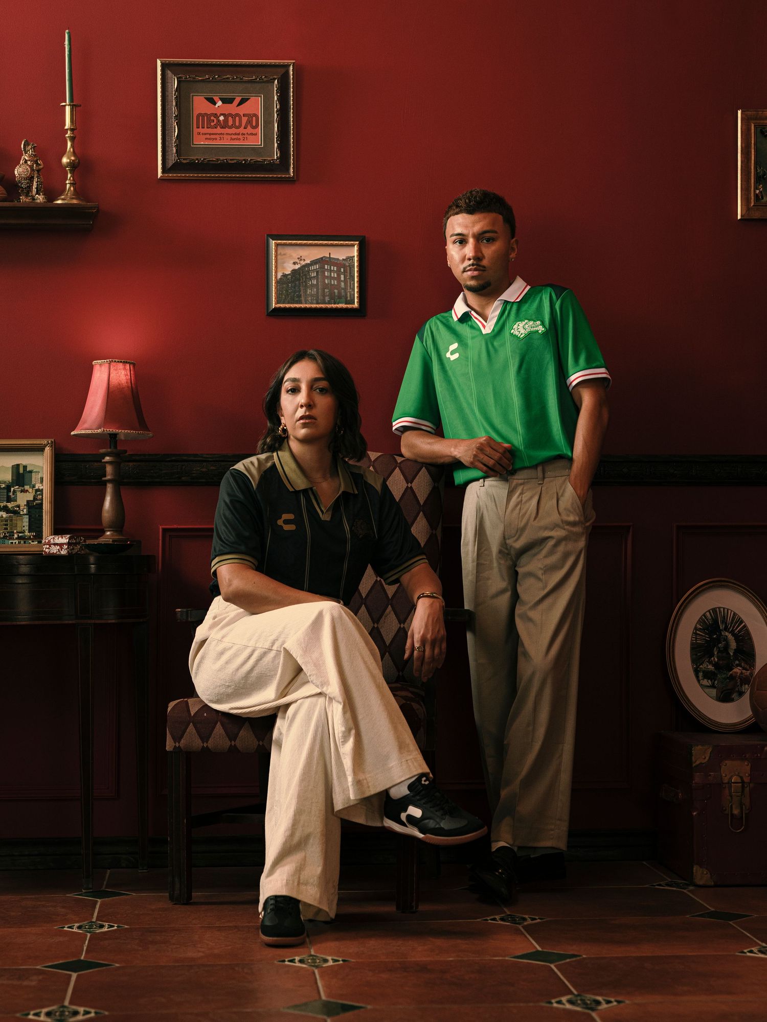 Two people pose in a red room wearing vintage-style green and dark sports jerseys with khaki pants.