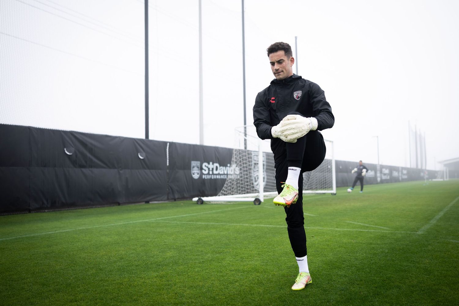Soccer goalkeeper warming up on a foggy training pitch, performing high knee drills in black uniform.