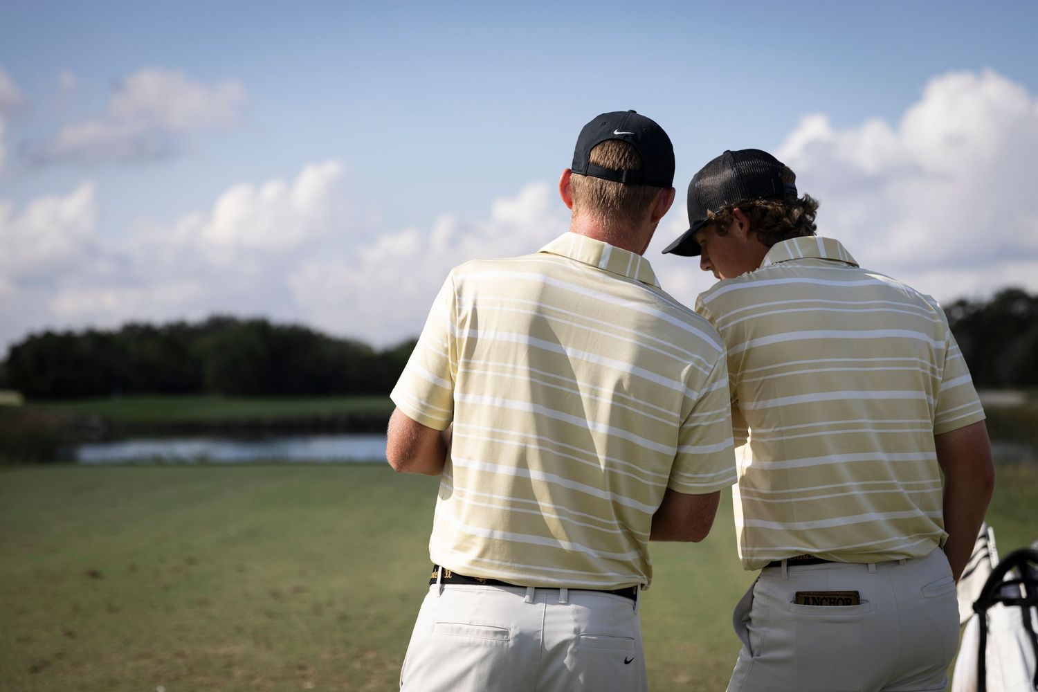 Two golfers in striped polo shirts stand together on course viewing the scenery.