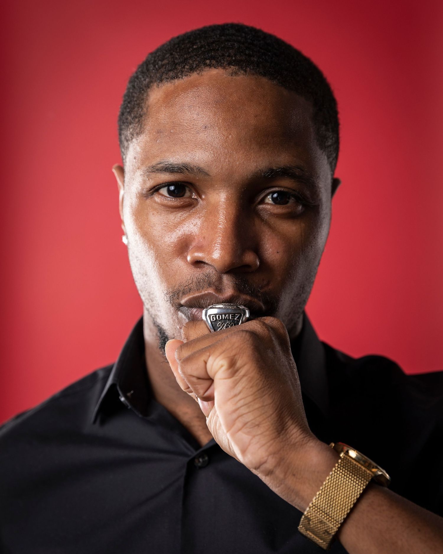 Athlete poses with championship ring against red background wearing black shirt and gold watch.