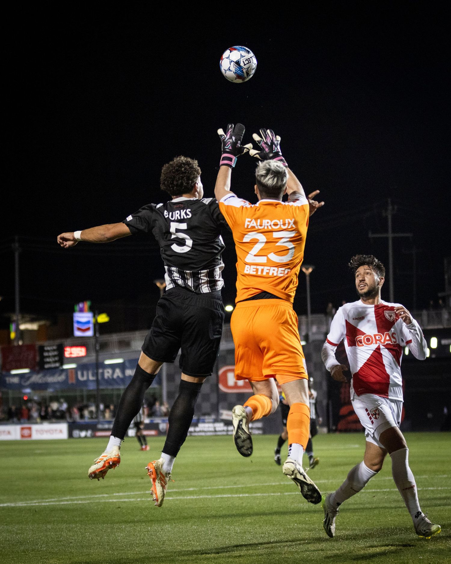 Three players leap for header ball with goalkeeper in orange jersey during night game.