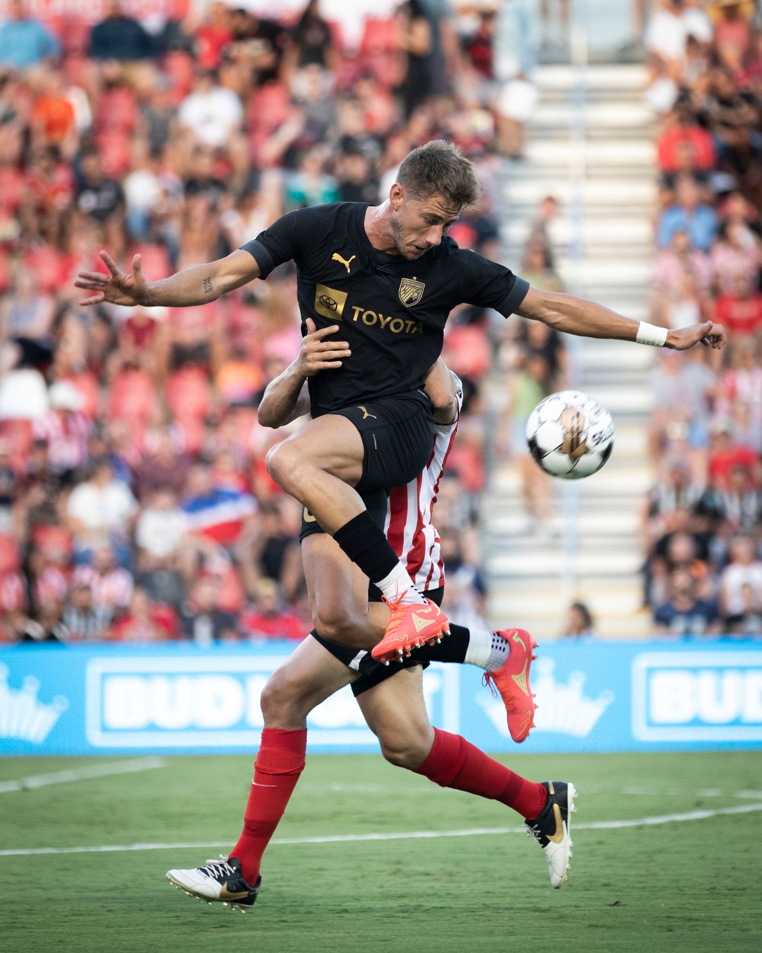 Soccer player in black jersey leaps over defender in intense aerial challenge.