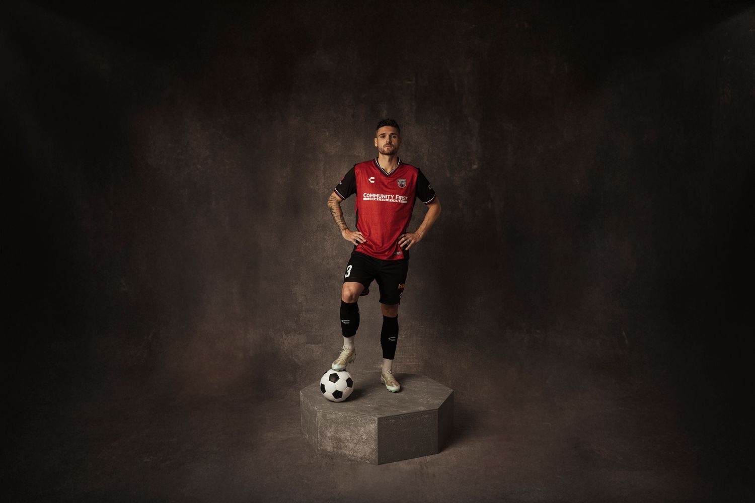Soccer player in red jersey stands confidently on a pedestal with a ball at their feet against a dark backdrop.