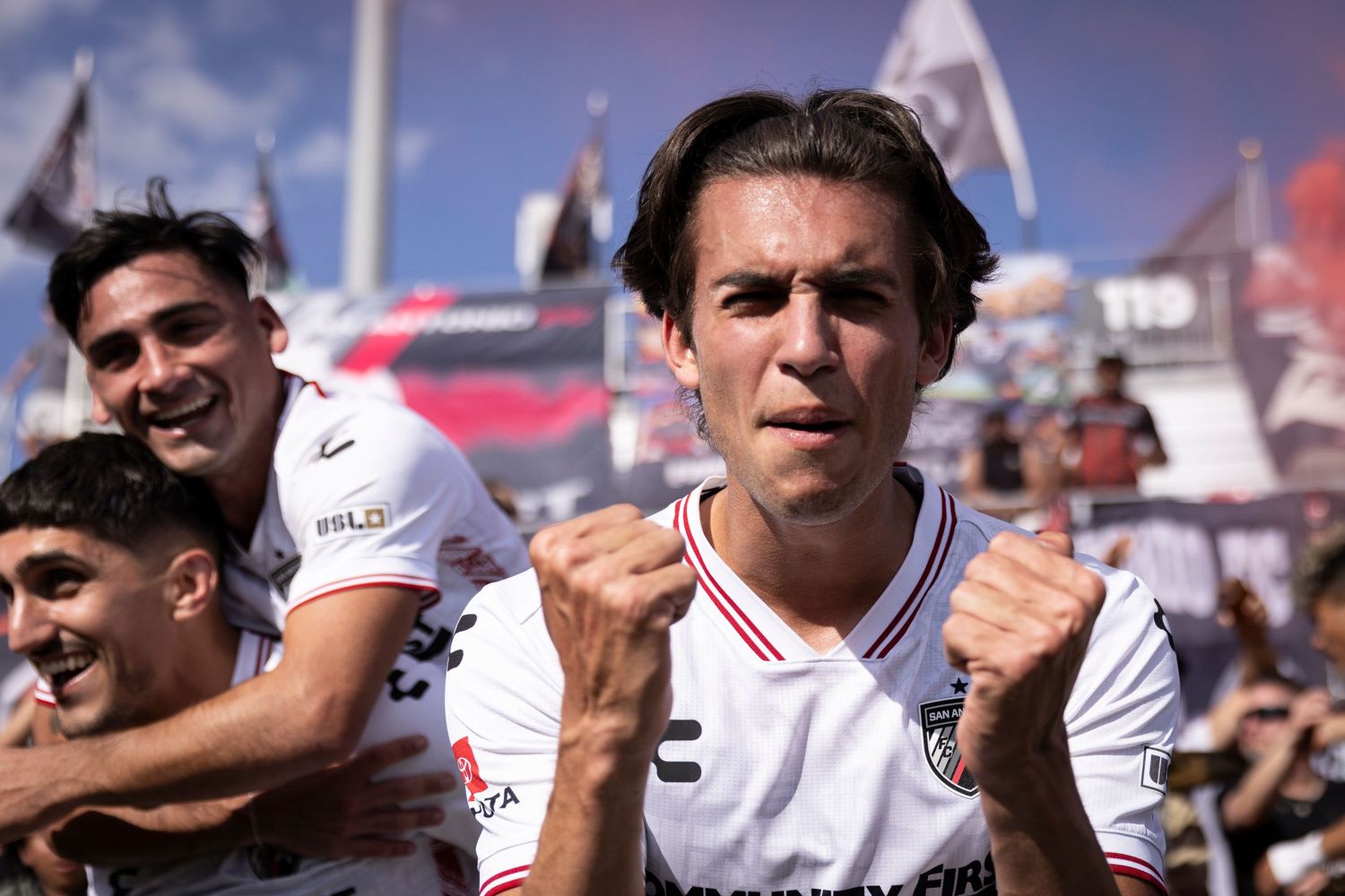 Soccer player celebrates a goal with fist pump as teammates embrace him from behind.