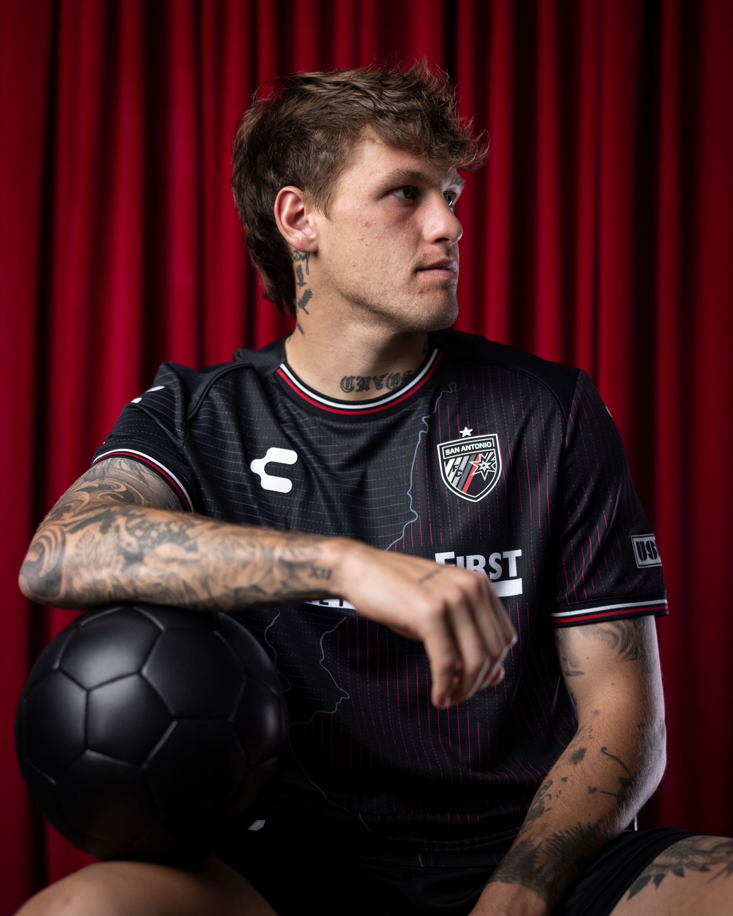 Soccer player in black Community First jersey poses with ball against red curtain.