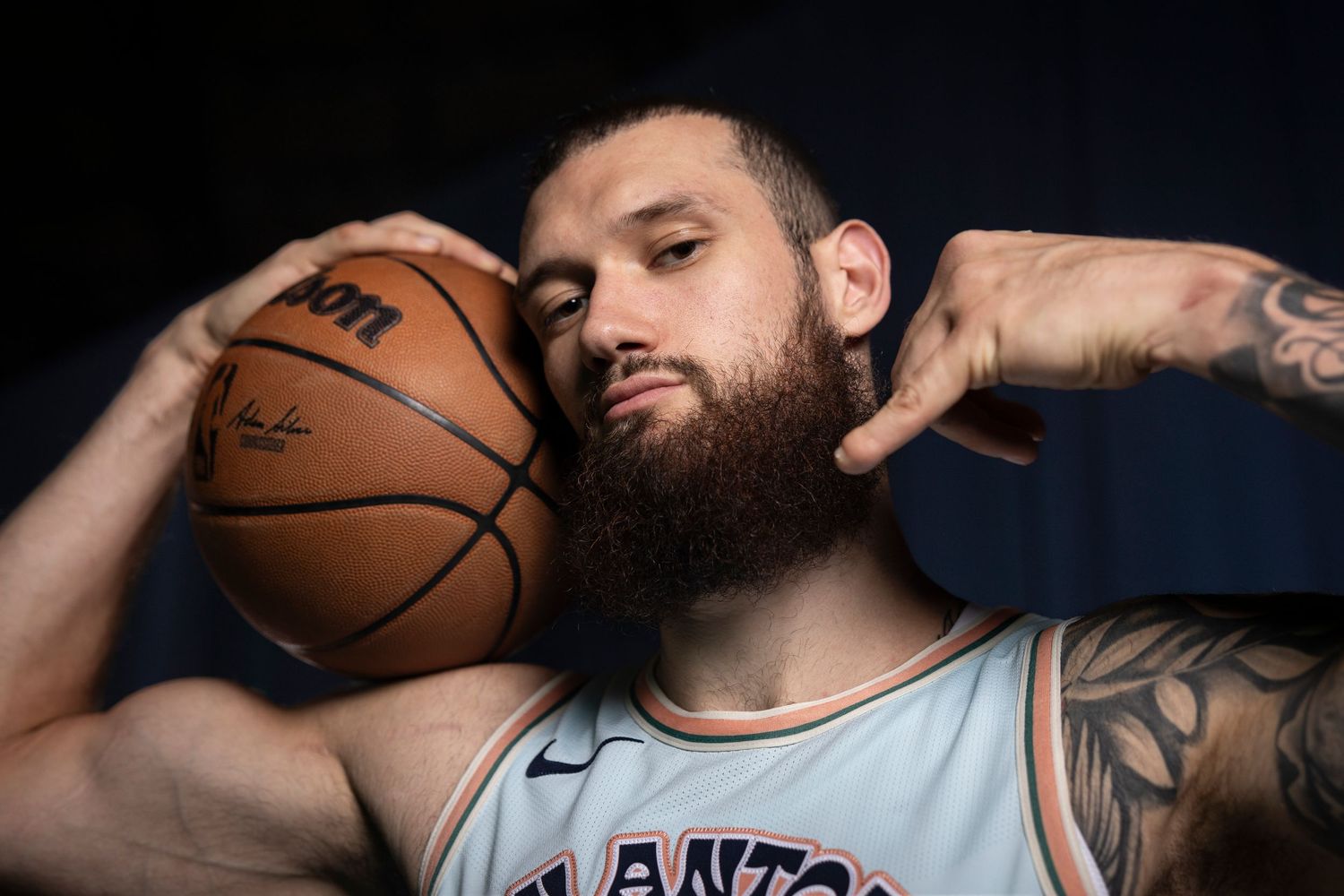 Professional basketball player holds a ball while showing off arm tattoos in dramatic lighting.