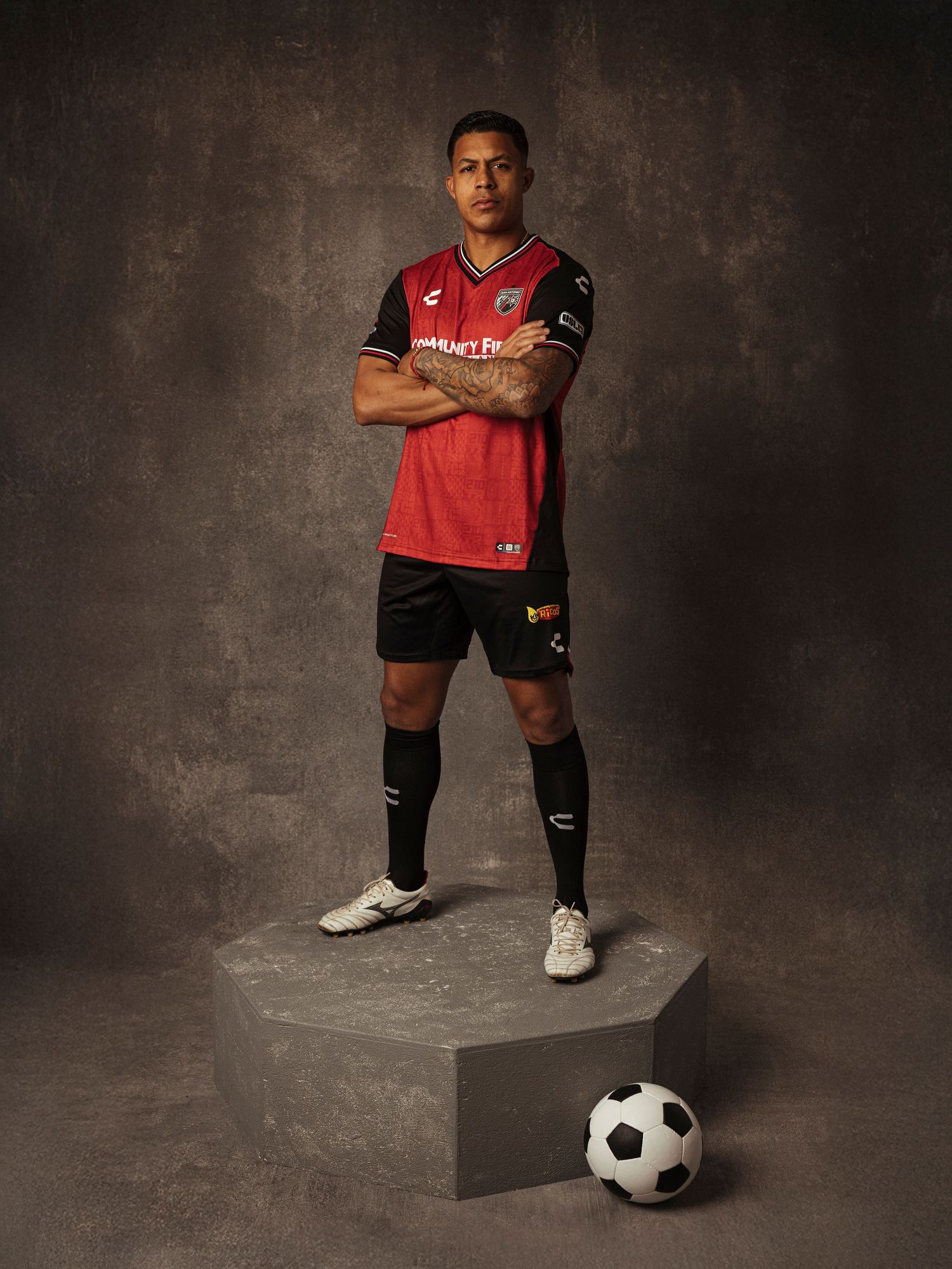 Soccer player in red and black Charly kit stands on platform with ball, arms crossed, studio portrait.