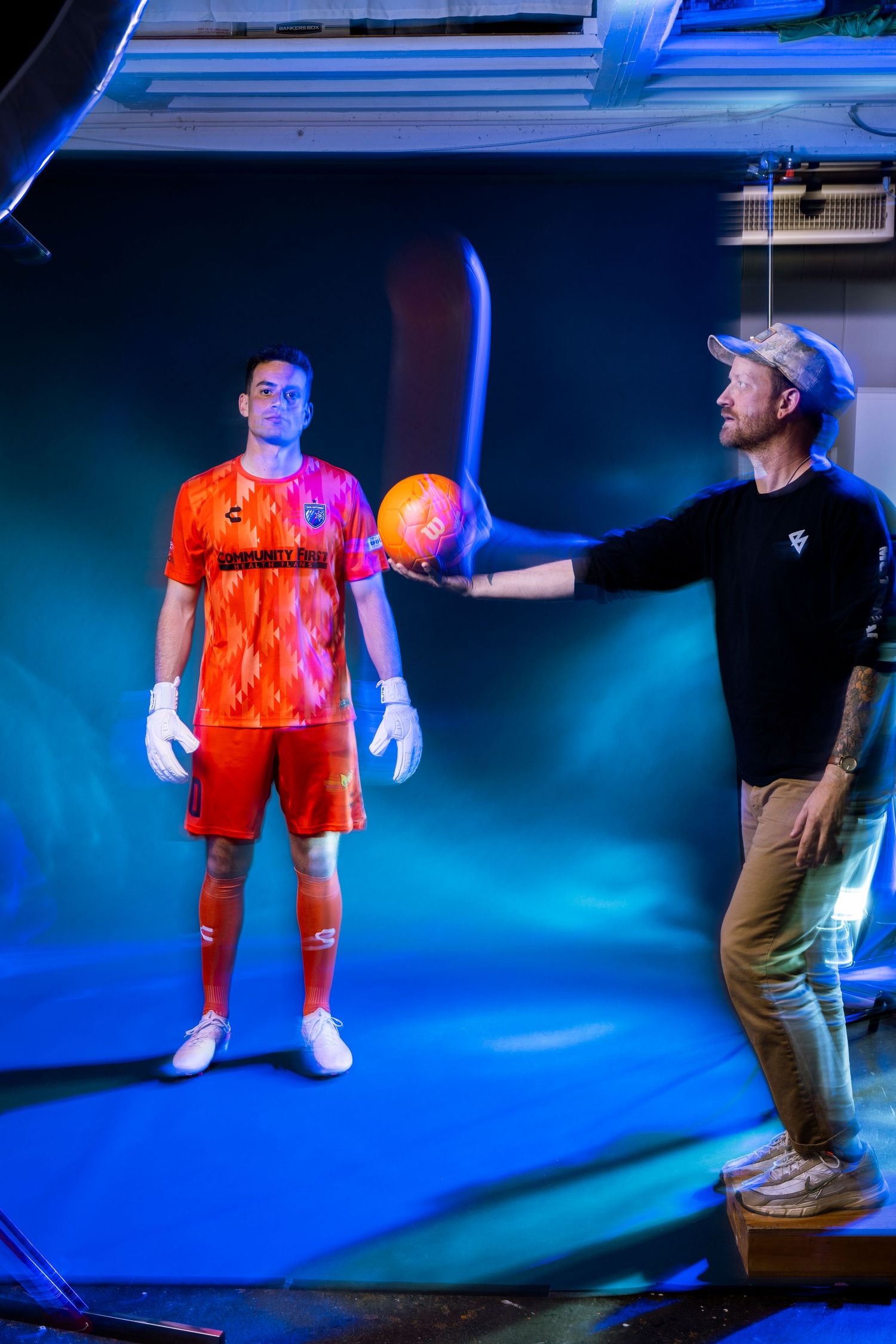 Soccer player in orange kit poses with ball held by photographer during professional photo shoot.
