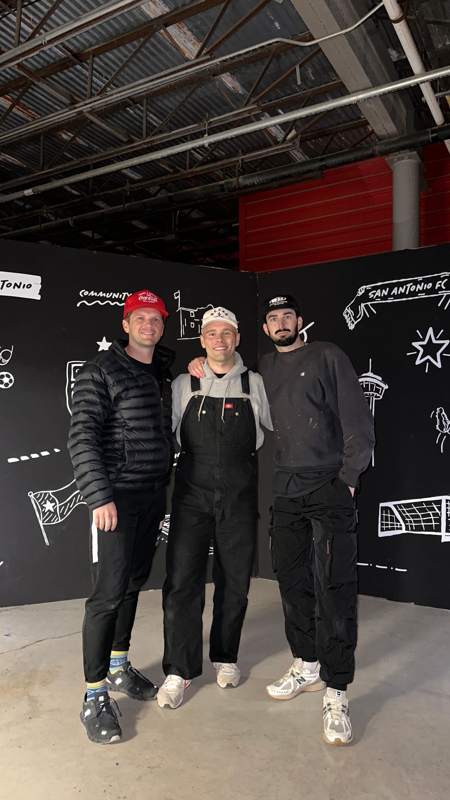 Three people stand together in front of a black wall with white chalk-style drawings in what appears to be an indoor venue.