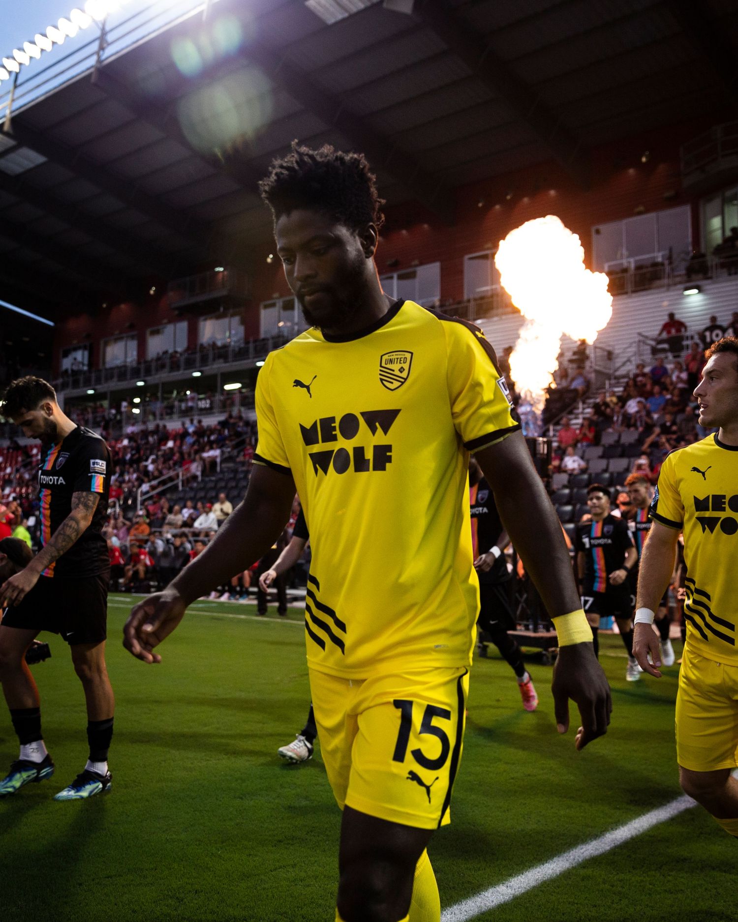 Soccer player in bright yellow jersey walks onto field with pyrotechnic effects in background.