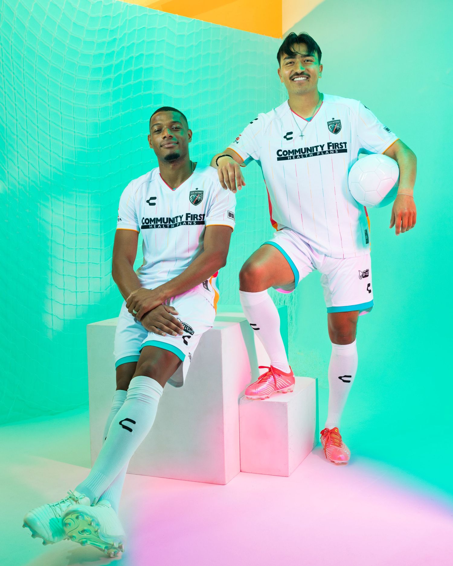 Two soccer players pose in white uniforms against colorful gradient background.