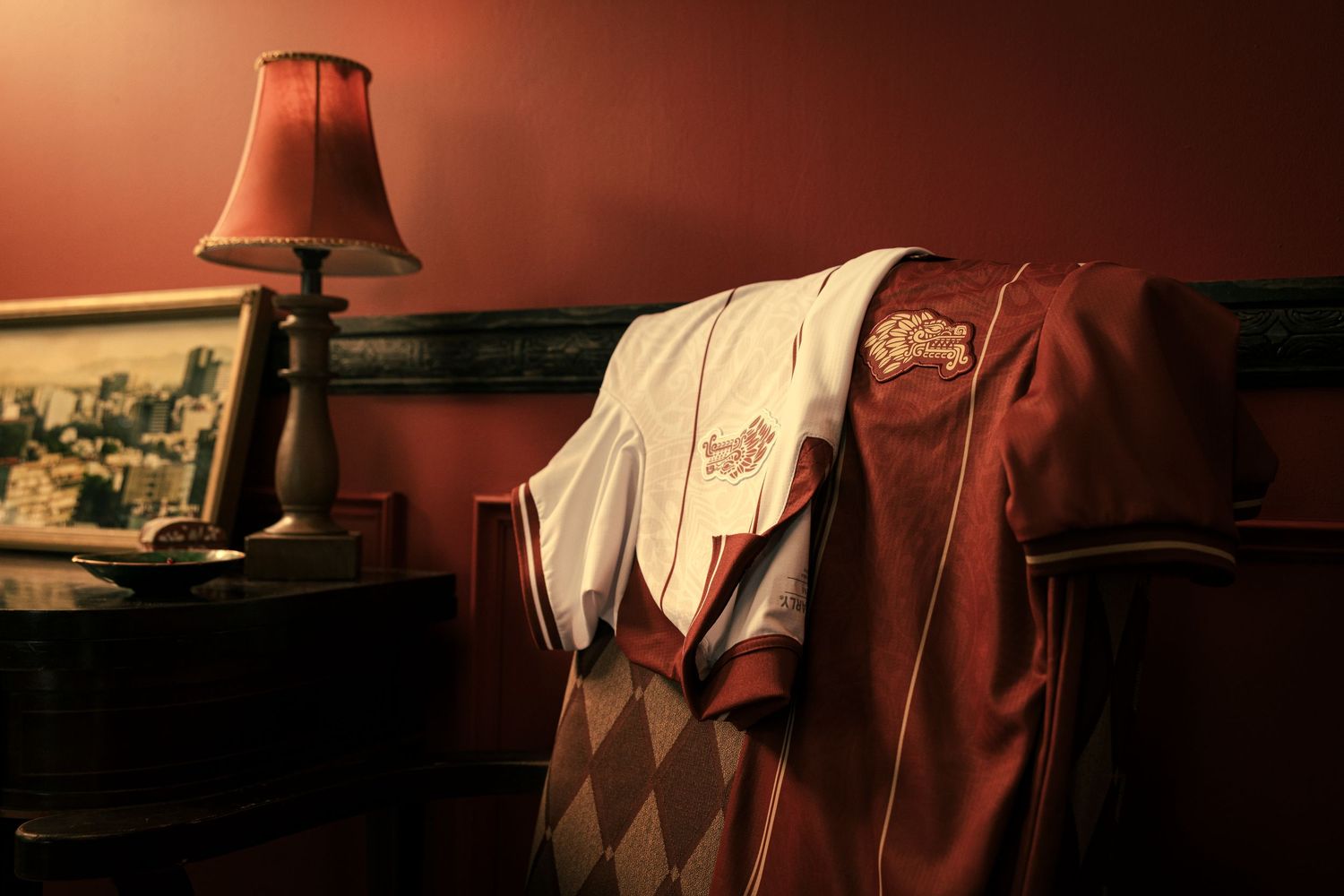 Maroon and white sports jacket draped over a chair beside a lamp on a nightstand in a warmly lit room.