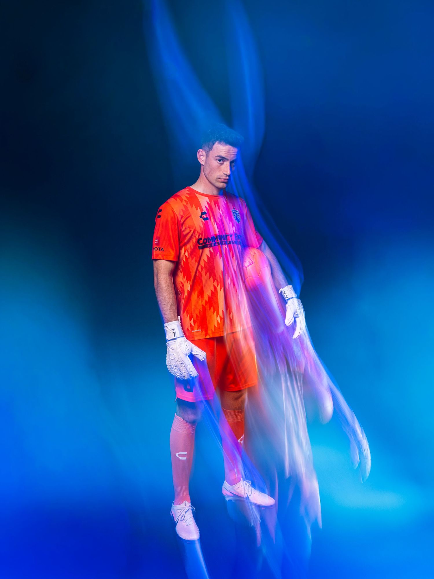 Soccer goalkeeper in vibrant orange kit with white gloves poses against a dramatic blue background with light streaks.