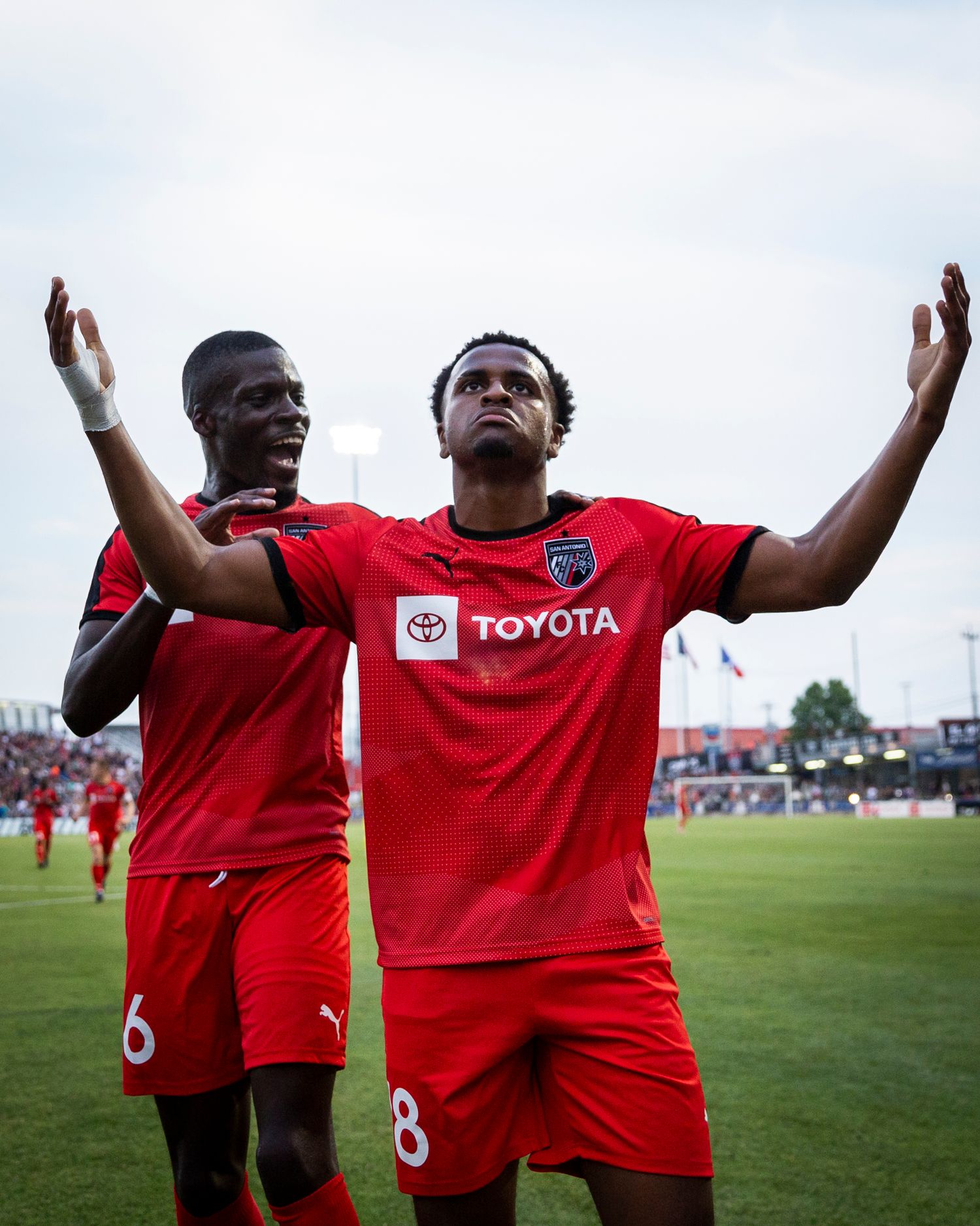 Two soccer players in red uniforms raise their arms in celebration on the pitch.