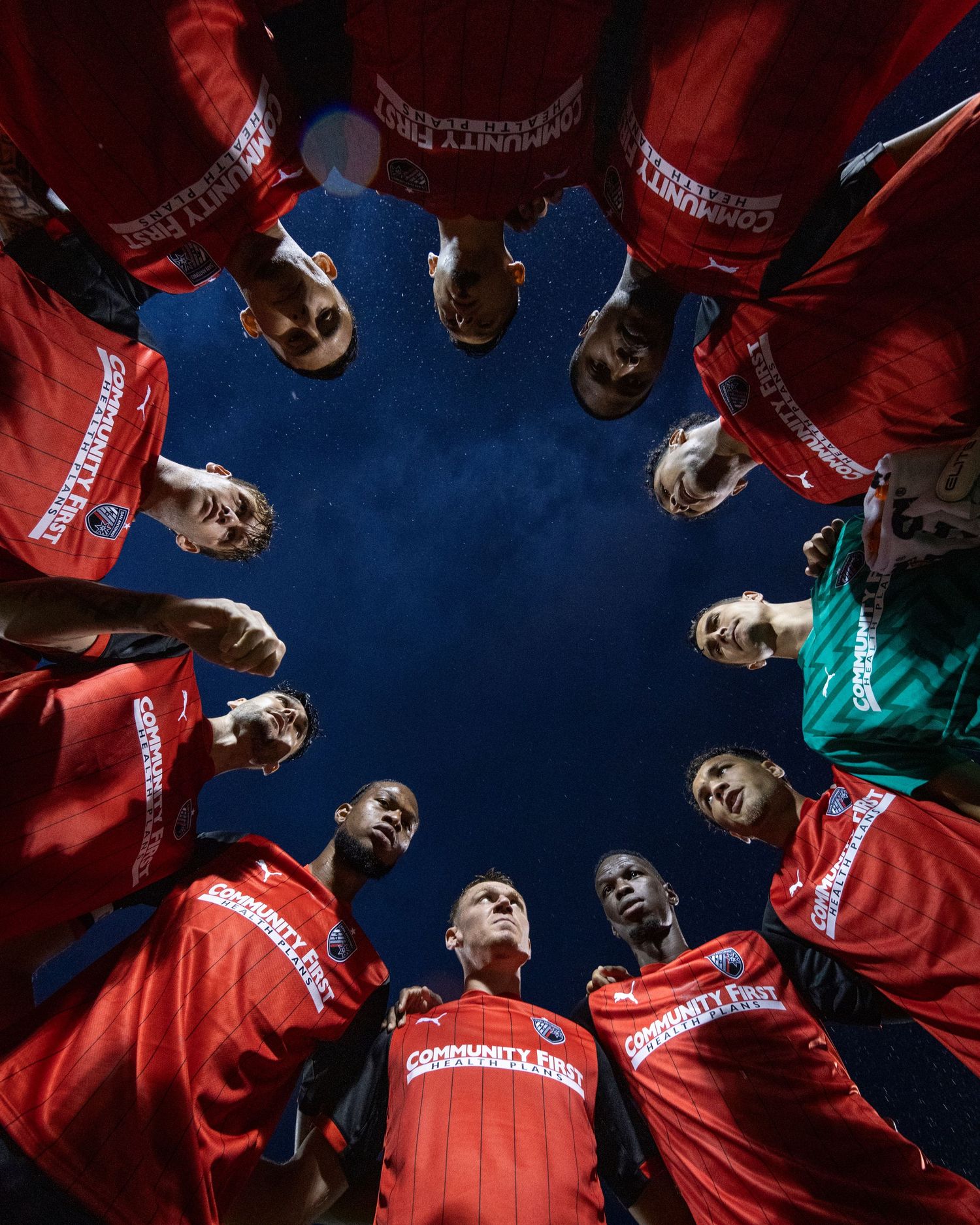 Soccer team huddles in circle formation wearing red jerseys against blue sky background.