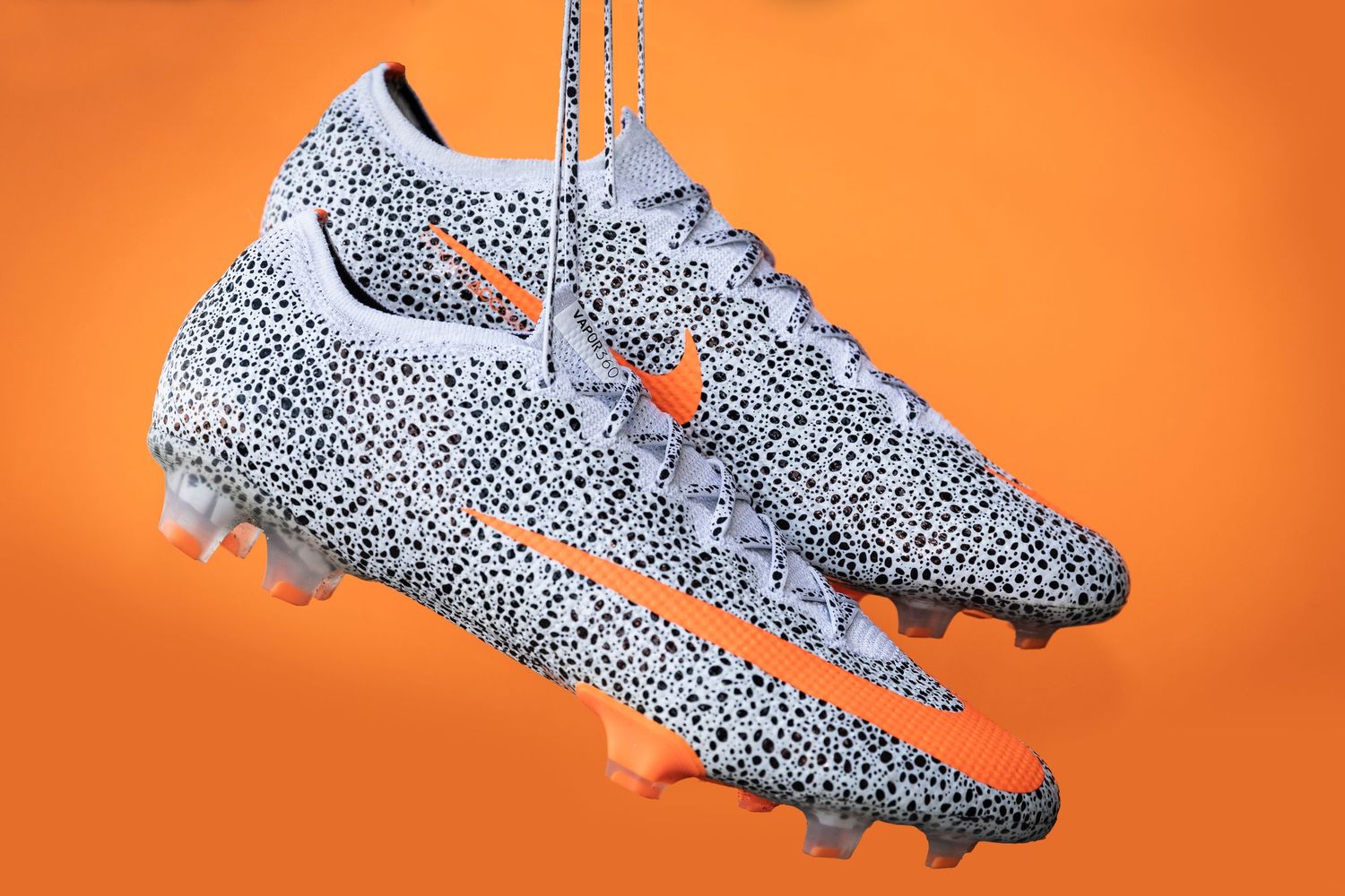 White and black spotted soccer cleats displayed on vibrant orange background.