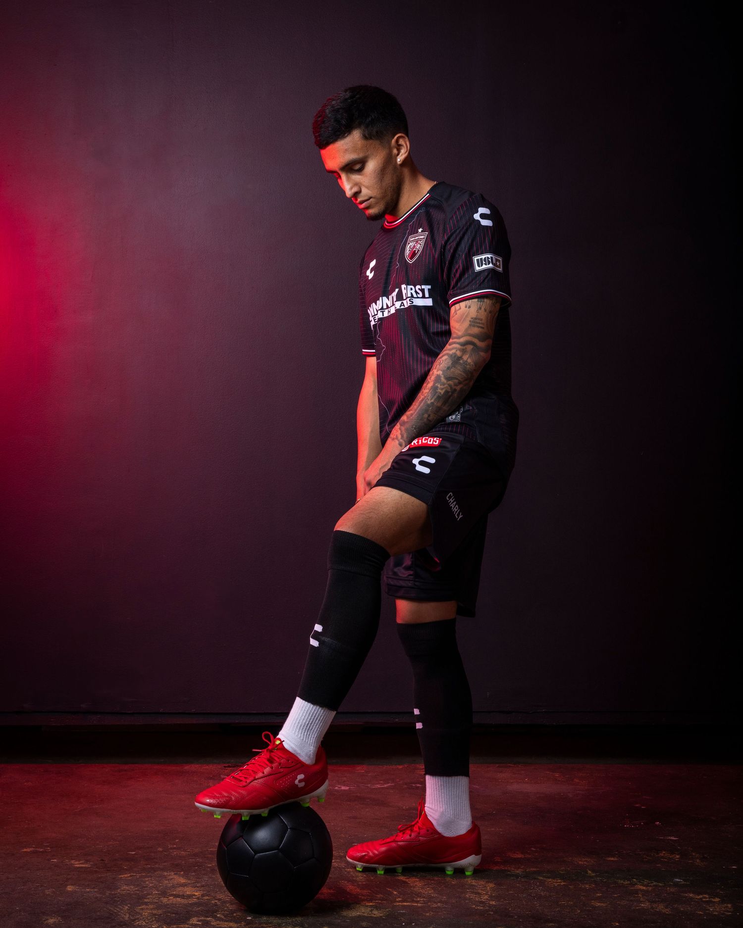 Soccer player in dark uniform poses with ball in dramatic red-lit studio setting with athletic stance and red cleats.