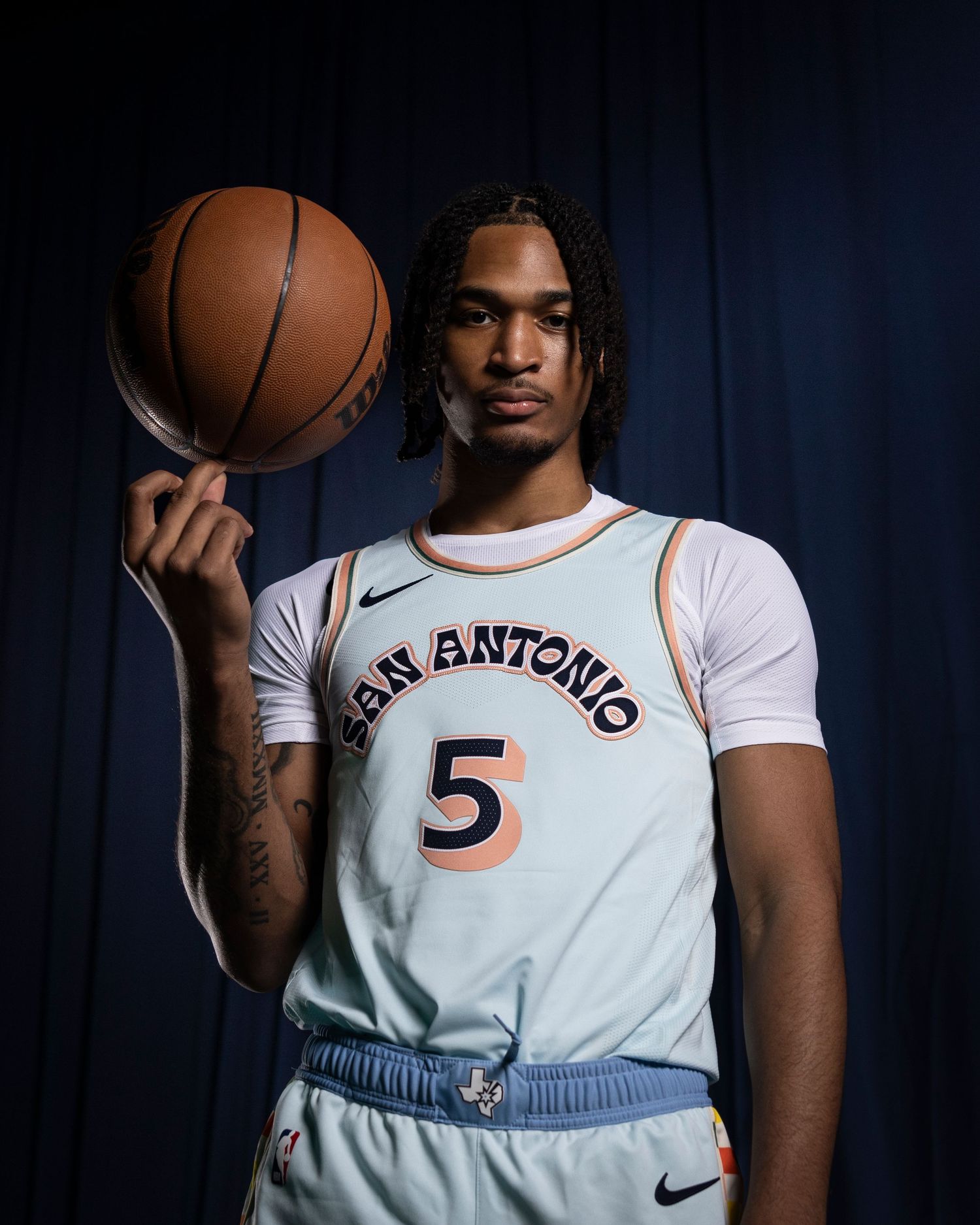 Basketball player in San Antonio number 5 jersey holds ball against dark background.