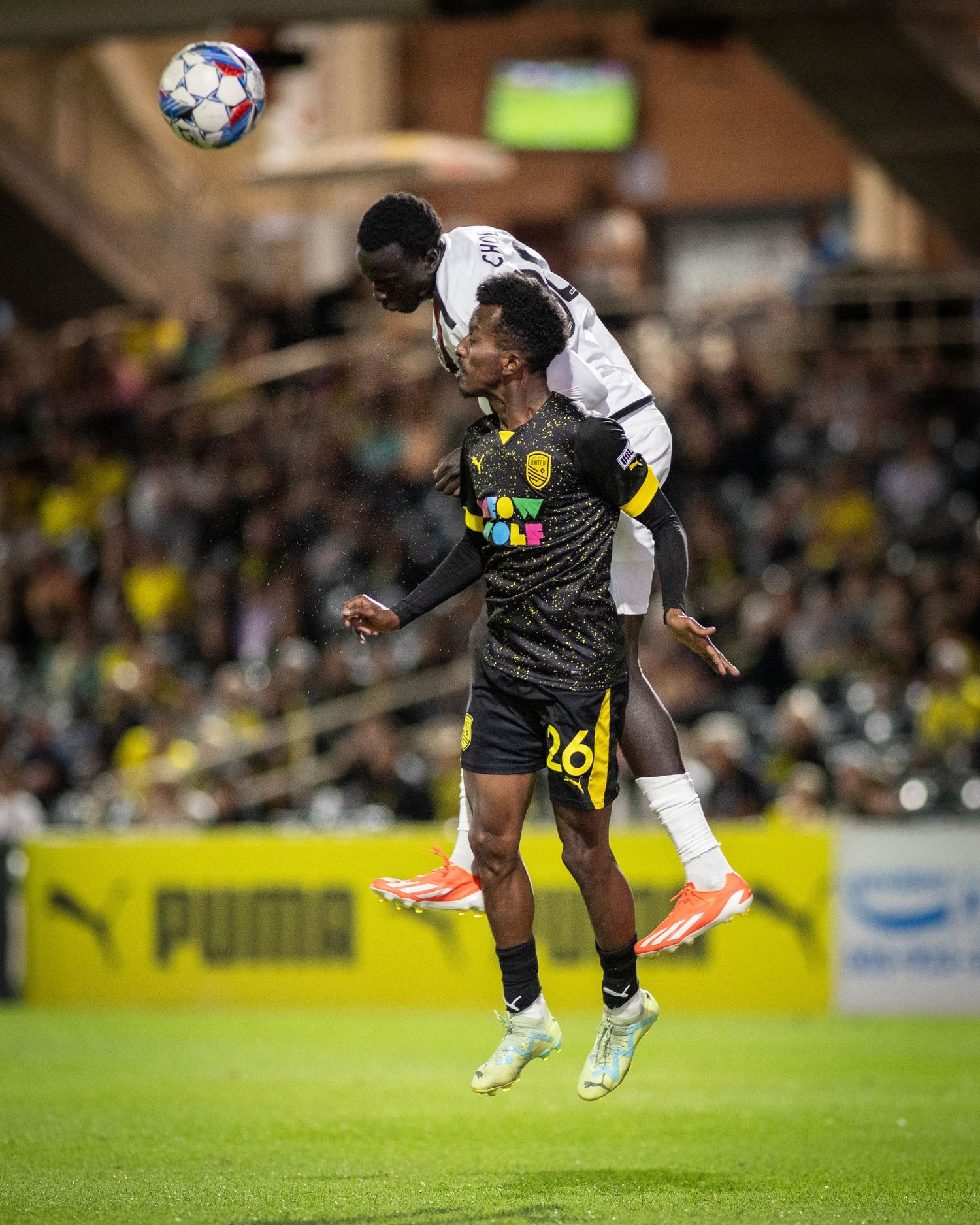 Two soccer players leap high in the air to contest a header during a night game.