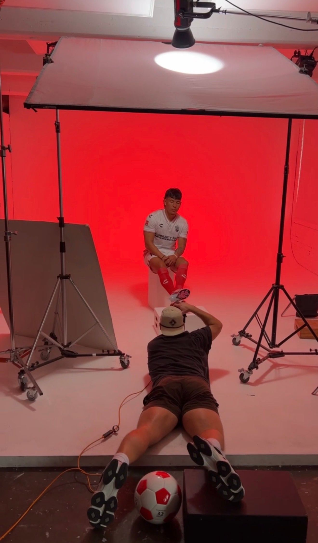 Two soccer players in a photography studio with red backdrop, one posing while another lies on floor with football.