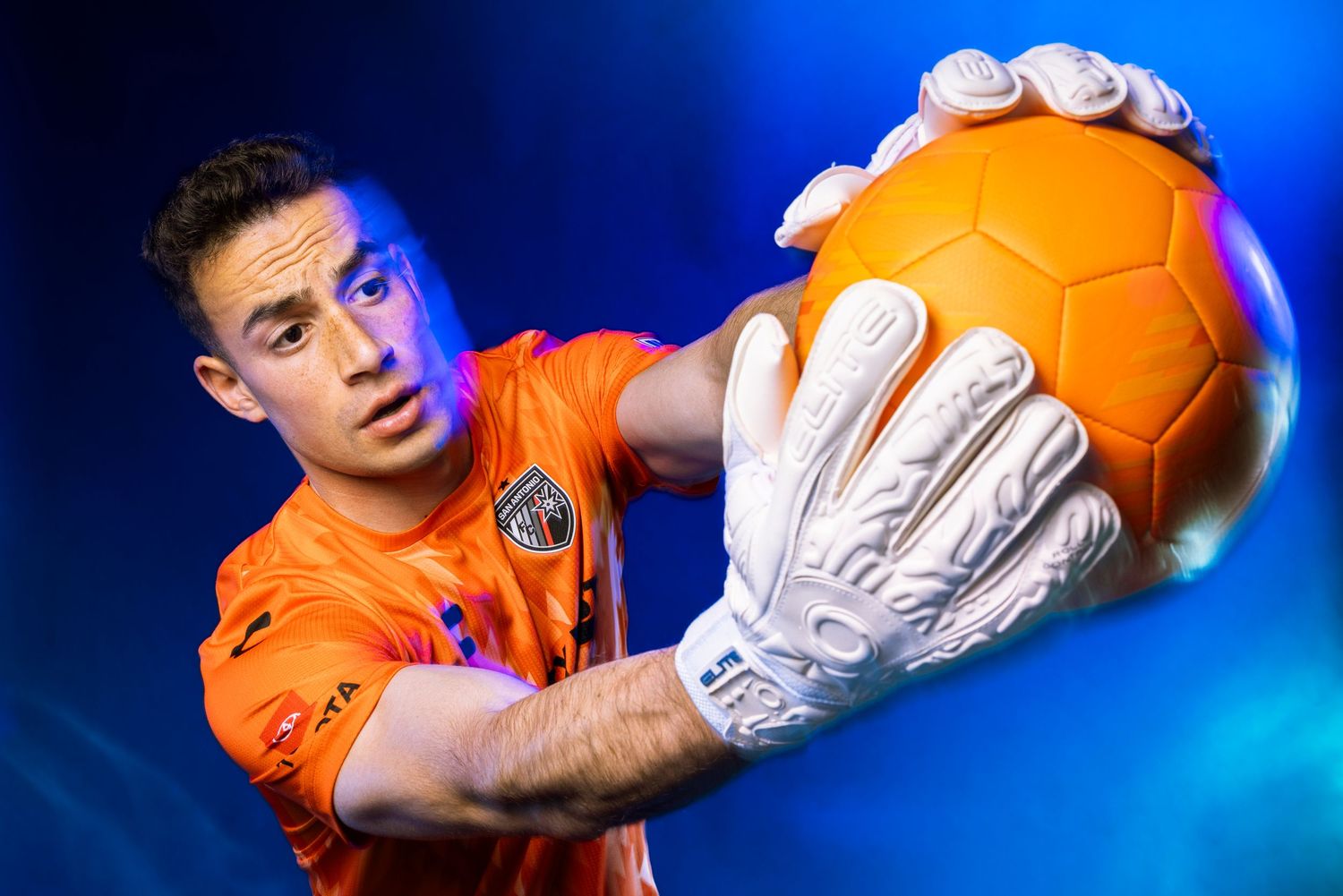 Male soccer goalkeeper in orange kit and white gloves holds an orange ball in a studio portrait shot.