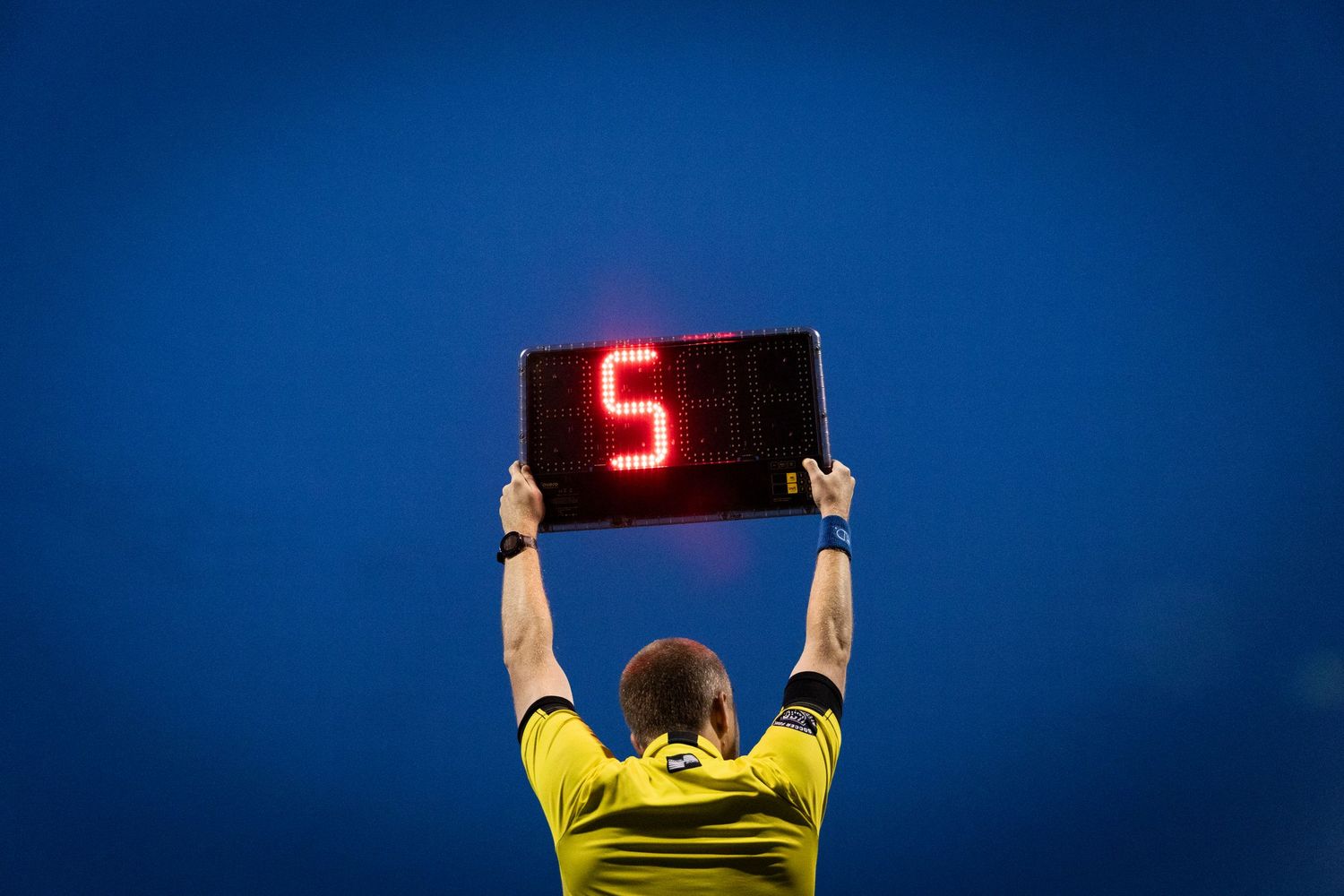 A referee in yellow holds up a digital substitution board showing number 5 against a dark blue sky.