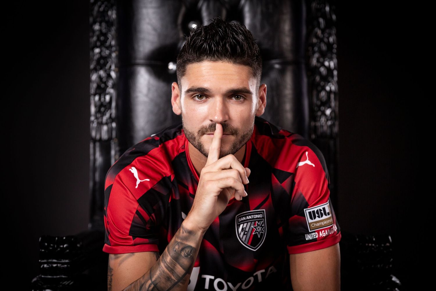 Soccer player in red jersey makes shushing gesture while posing against dark background.
