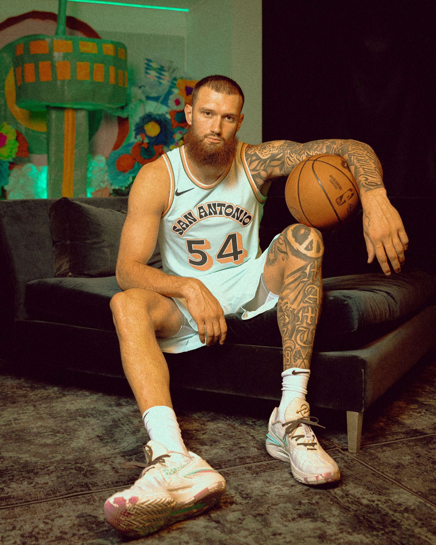 Basketball player in San Antonio jersey number 54 sits on couch holding ball.