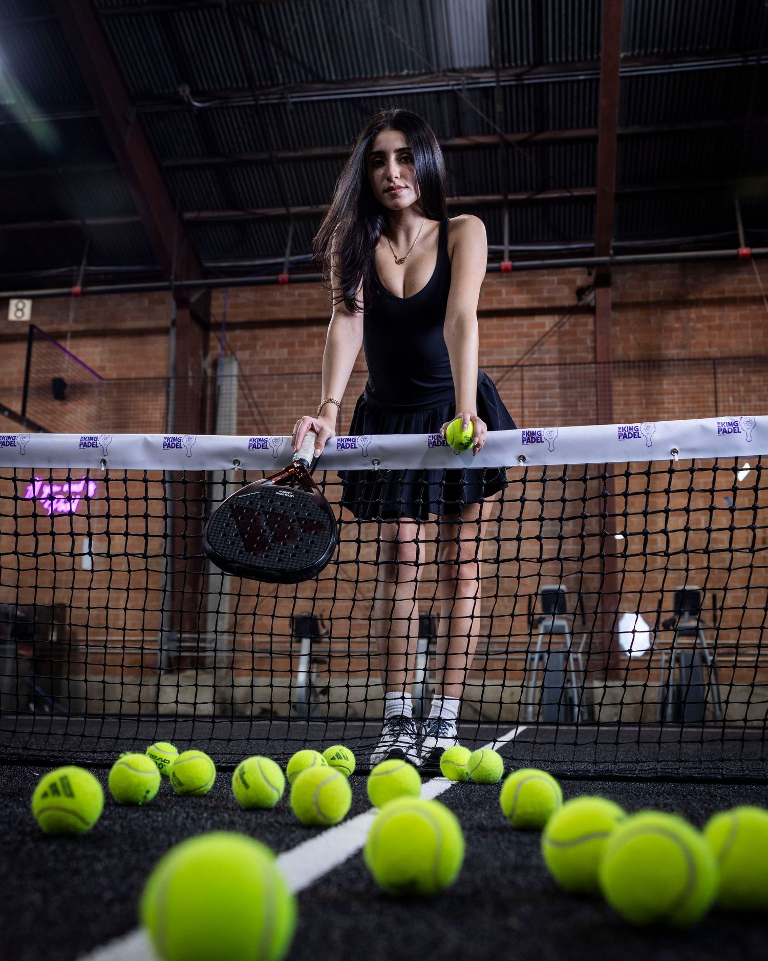 Athletic figure at pickleball net with scattered neon balls in dark indoor court setting.