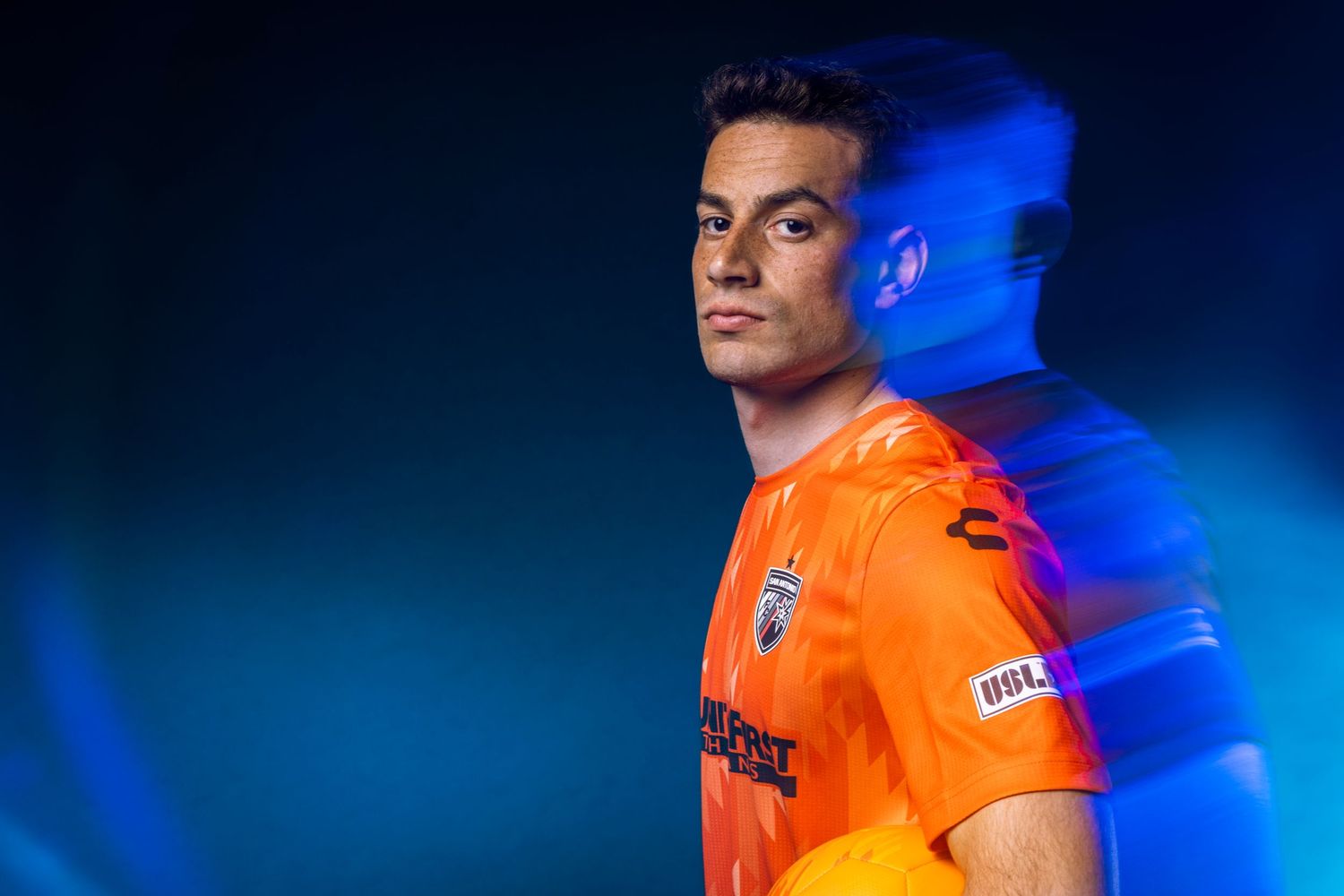 Soccer player in orange UFL jersey poses confidently against a vibrant blue light background.