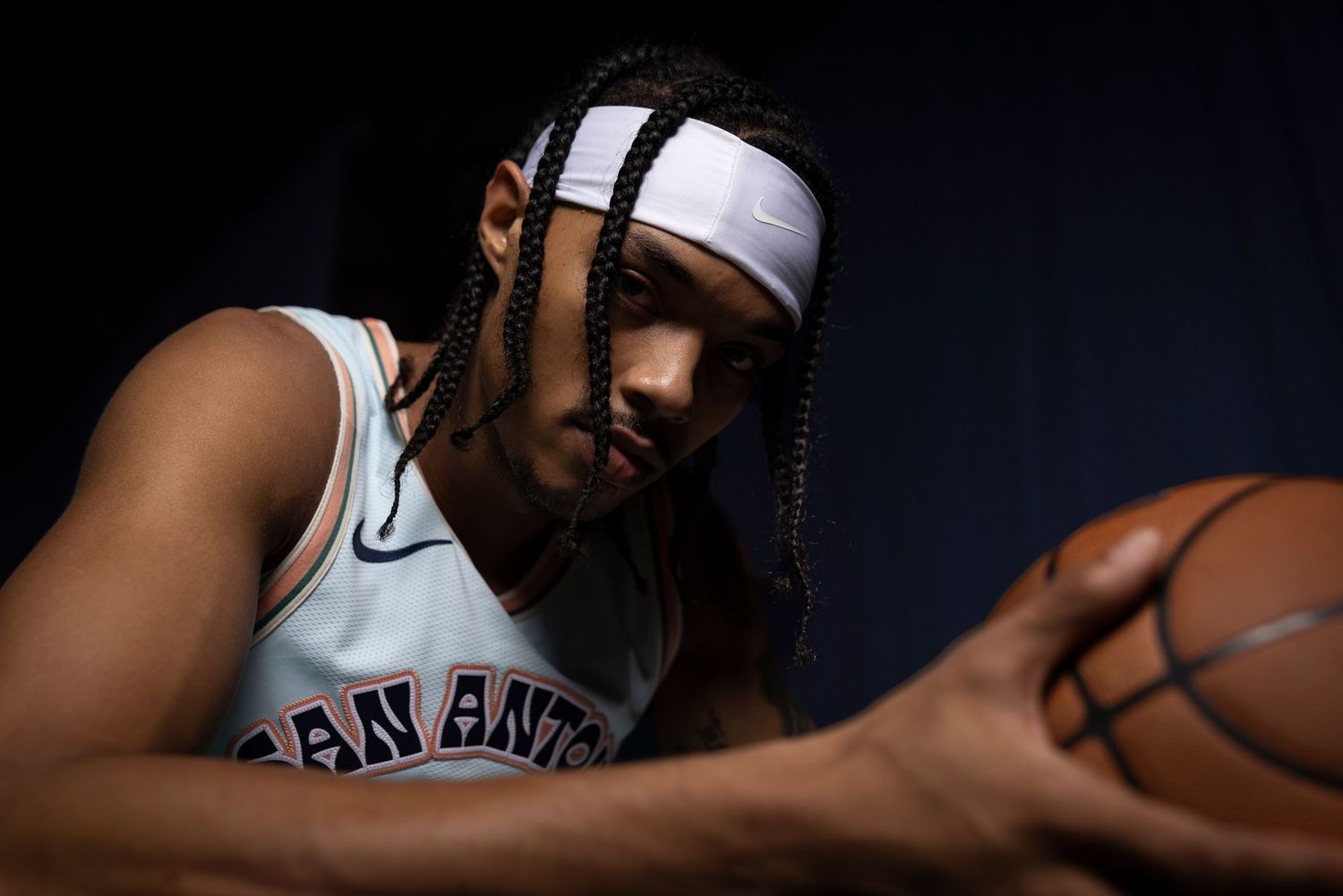 San Antonio Spurs player in Nike jersey holds basketball, wearing white headband with braided hair in dramatic studio lighting.
