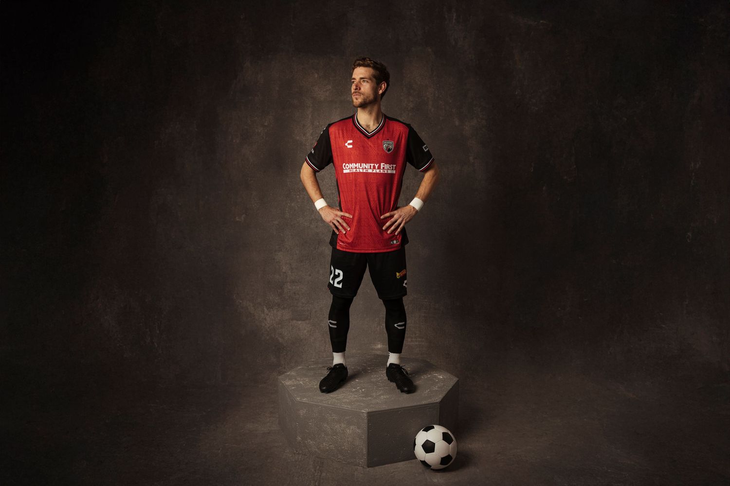 Soccer player in red and black uniform stands confidently with hands on hips, soccer ball beside him on studio backdrop.