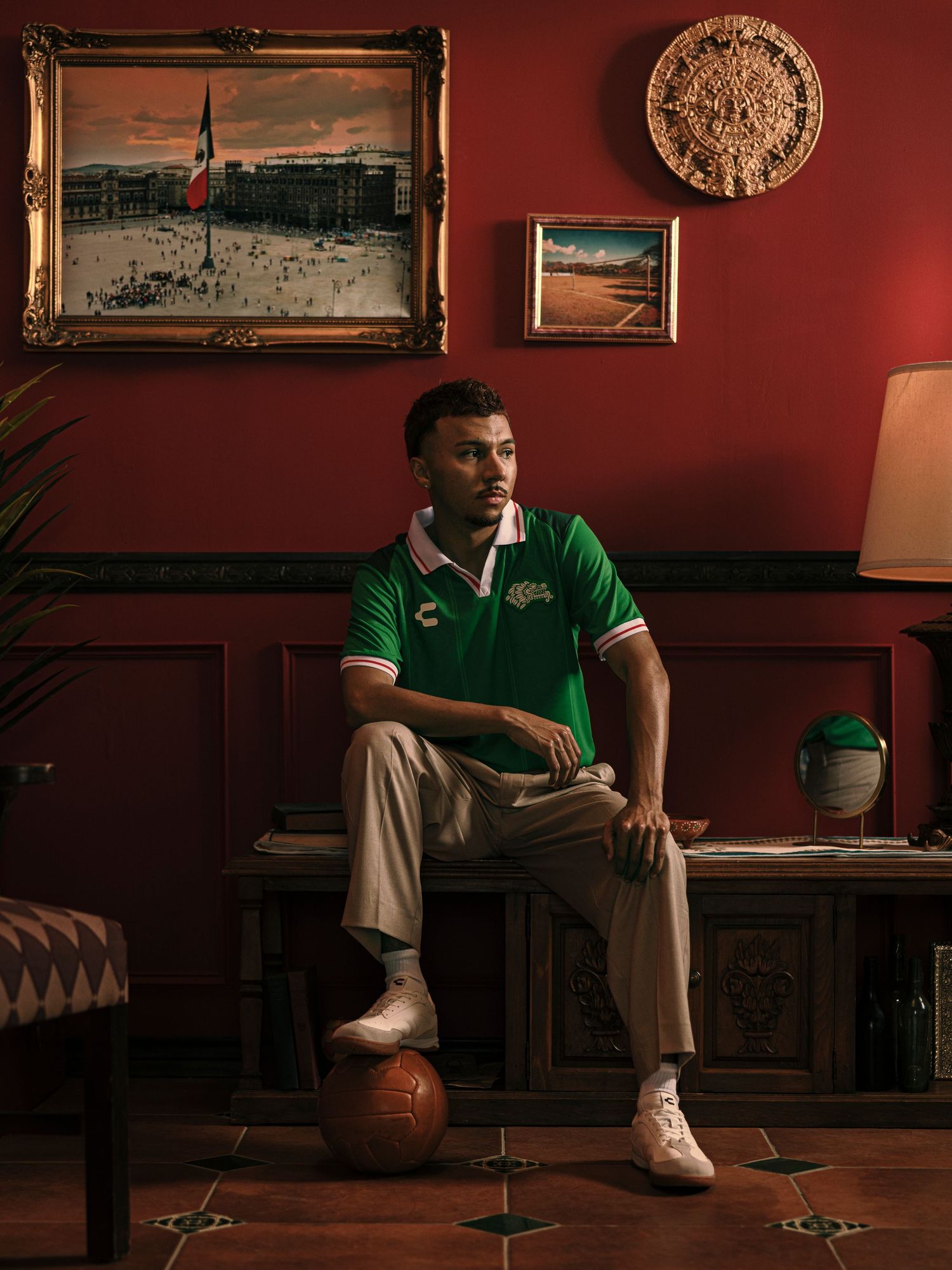 Man in green Mexico soccer jersey sits on a soccer ball in a red room with Mexican artwork on the walls.