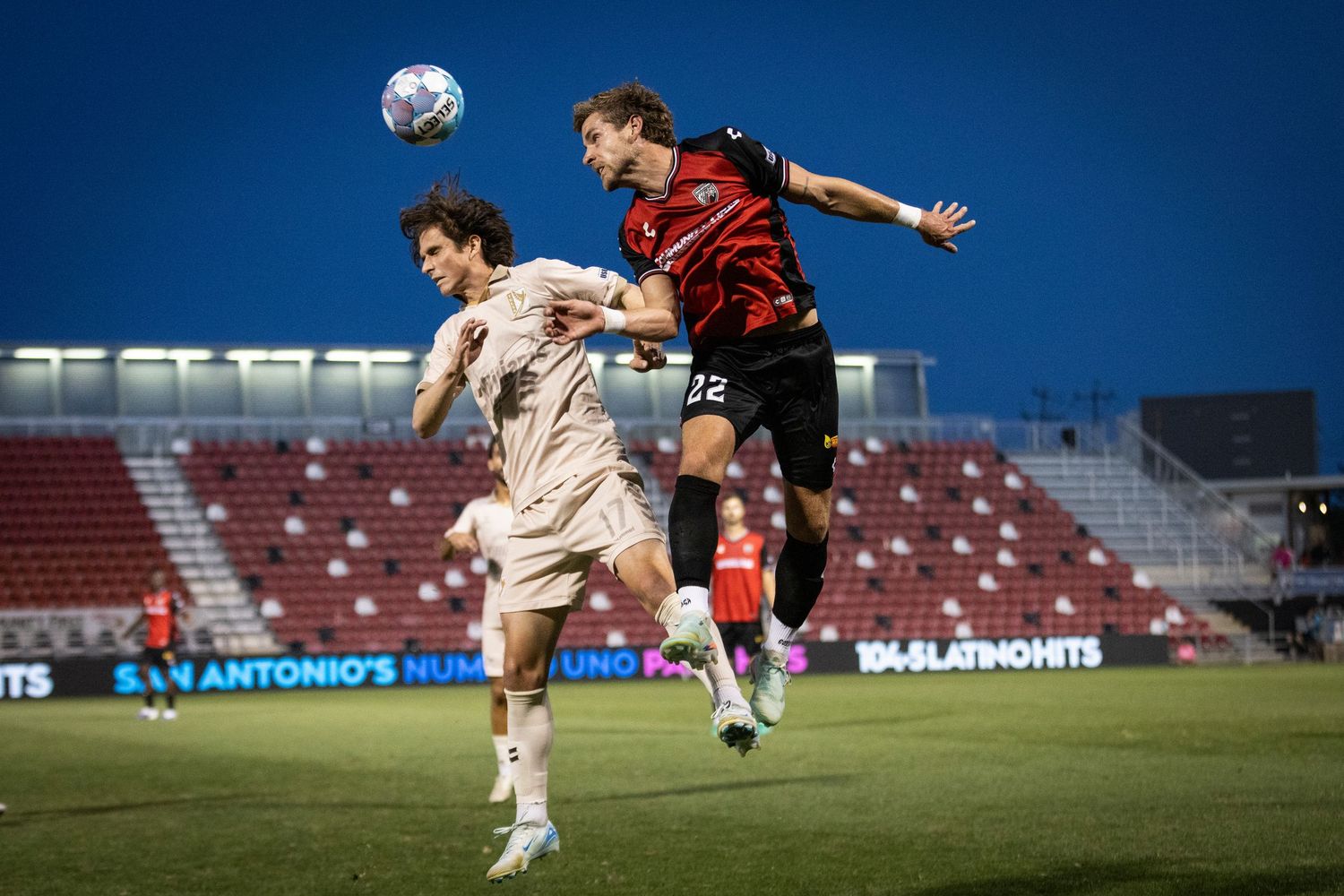 Two soccer players battle for a header during a night match at San Antonio FC stadium under bright floodlights.