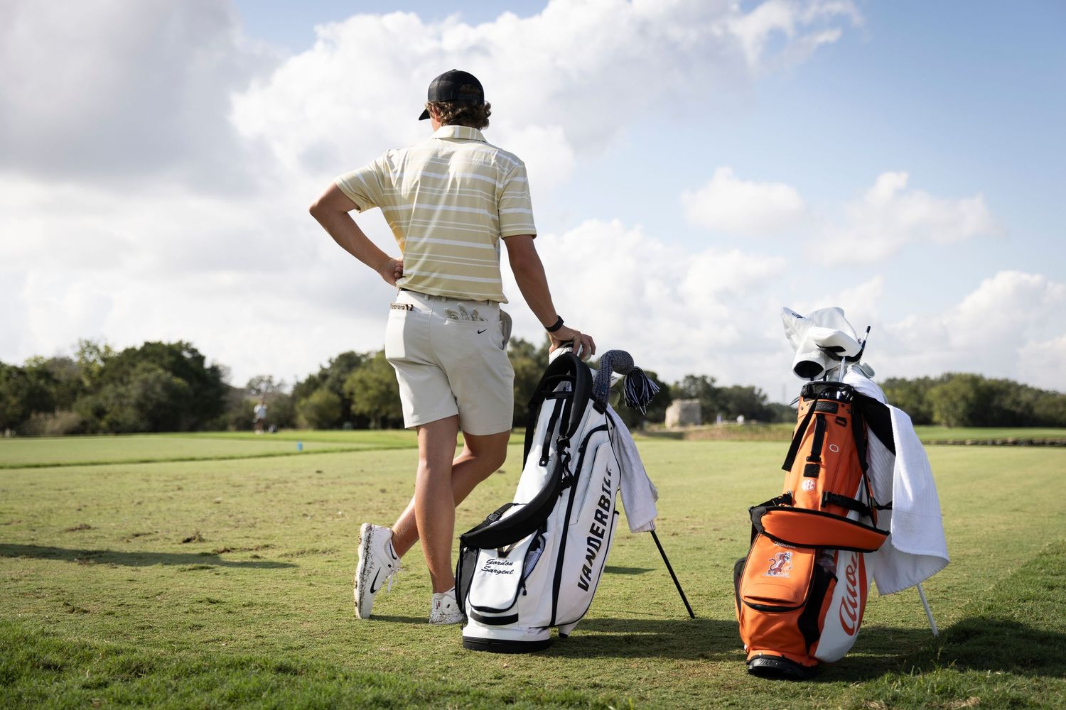 Golfer standing with golf bags on course under blue sky and white clouds.