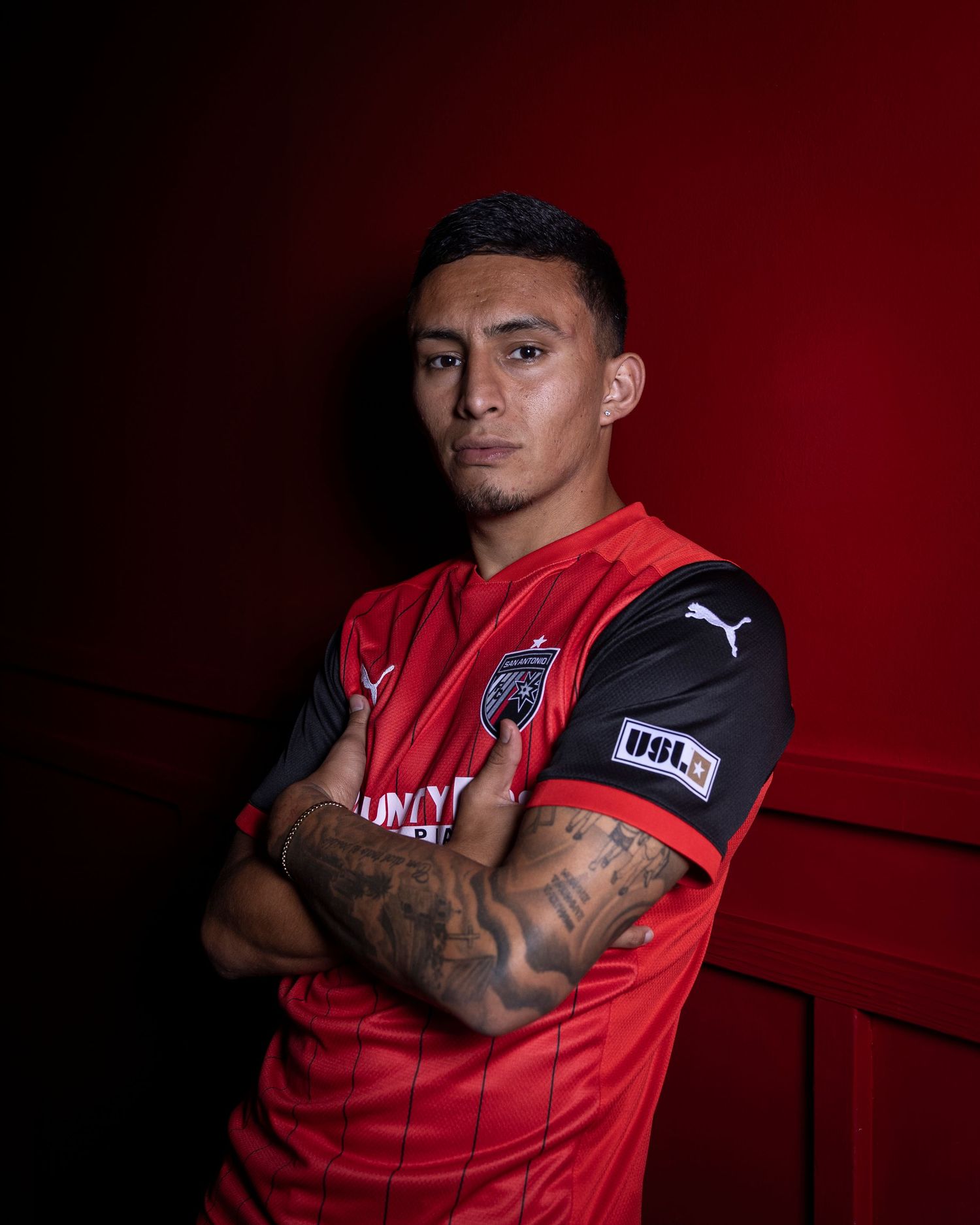 Professional soccer player poses in a red Puma jersey with tattooed arms crossed against dark background.