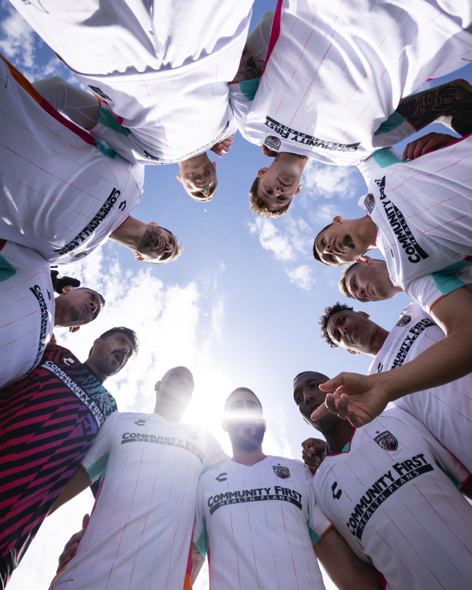 Soccer team huddles in a circle formation against bright sky background.