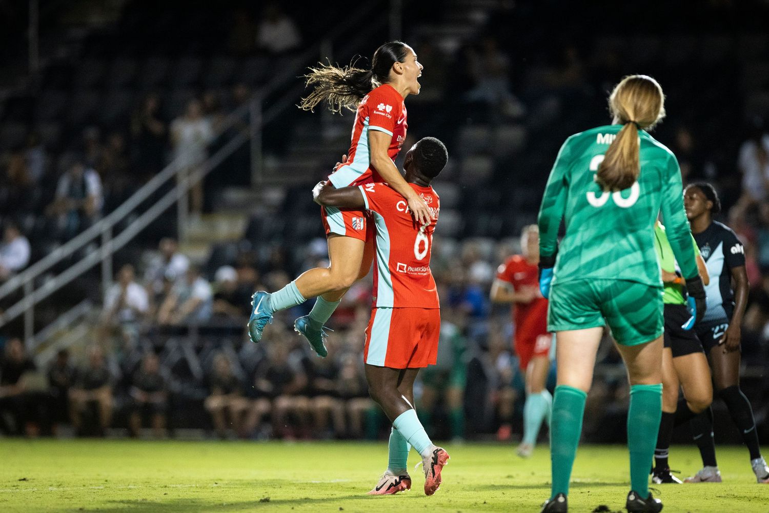 Players jump for header during soccer match with goalkeeper watching in background.