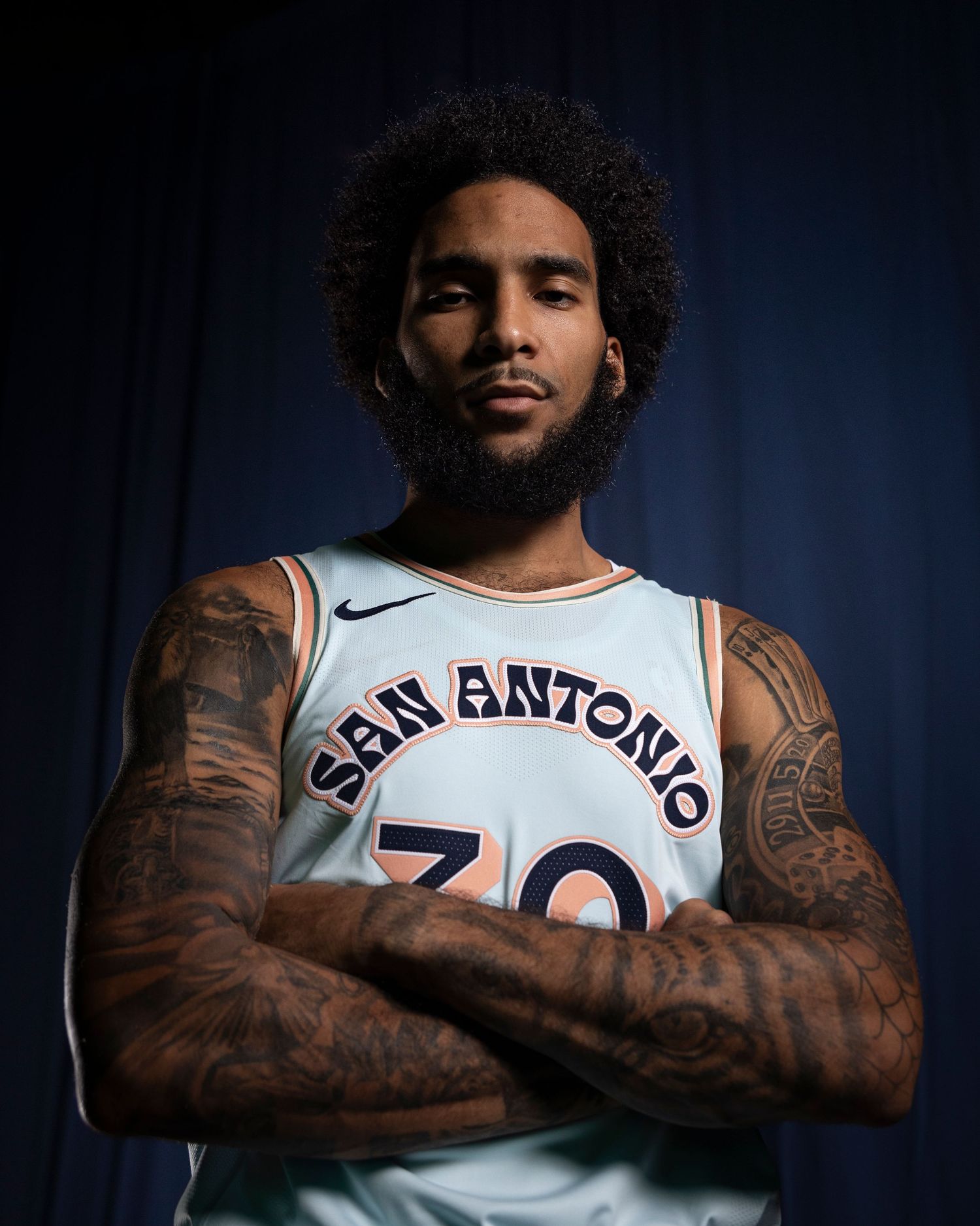Professional basketball player in San Antonio jersey shows off tattooed arms against dark background.