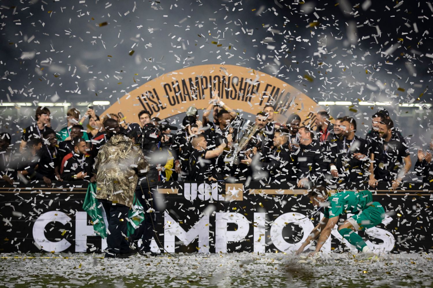 Championship celebration on field with confetti flying as team celebrates behind large CHAMPIONS sign.
